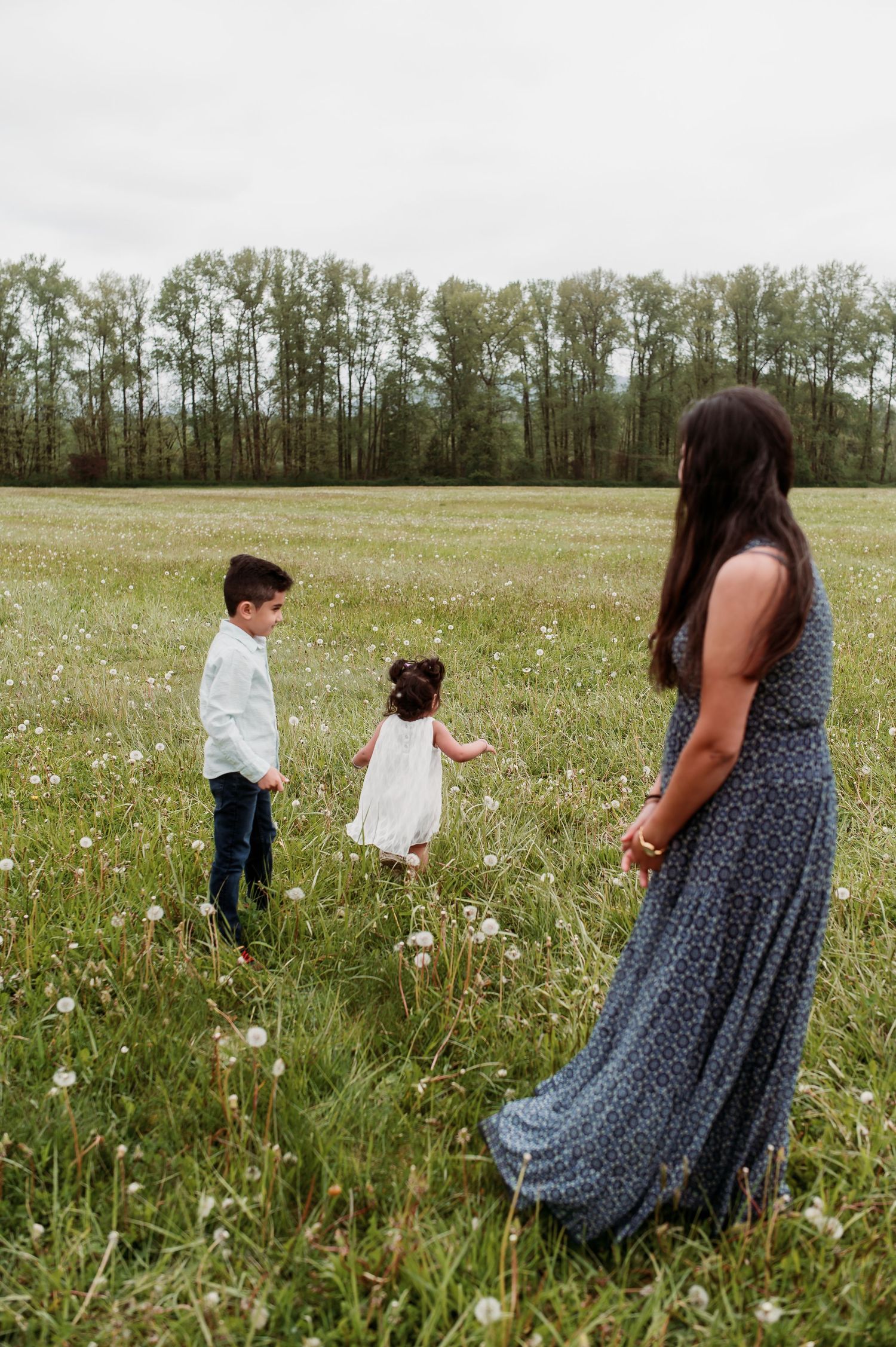People in a wildflower meadow with tall trees in background, woman wearing long blue floral dress stands near children.