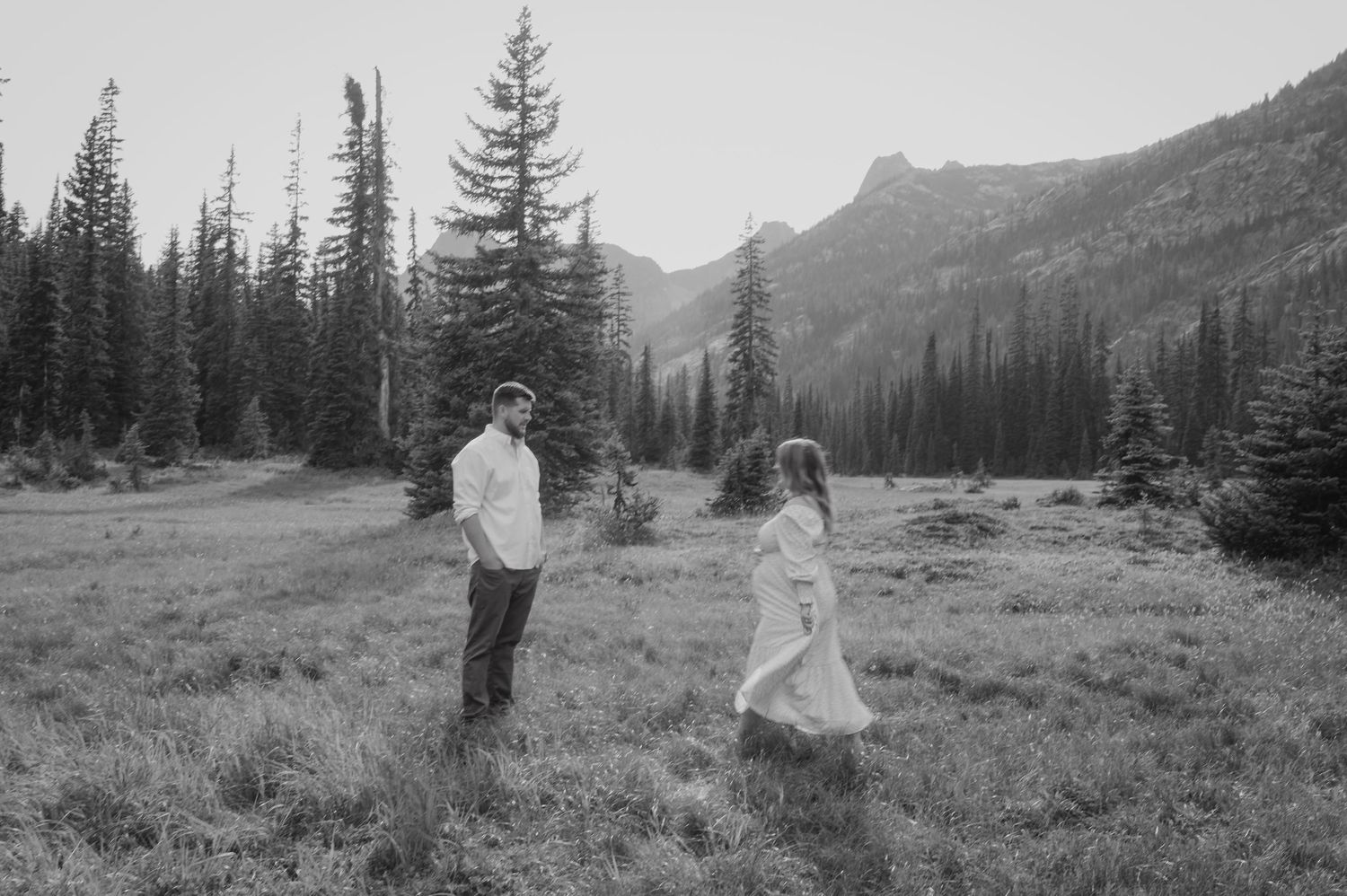 Black and white photograph of figures in a mountain meadow surrounded by towering evergreens and peaks.