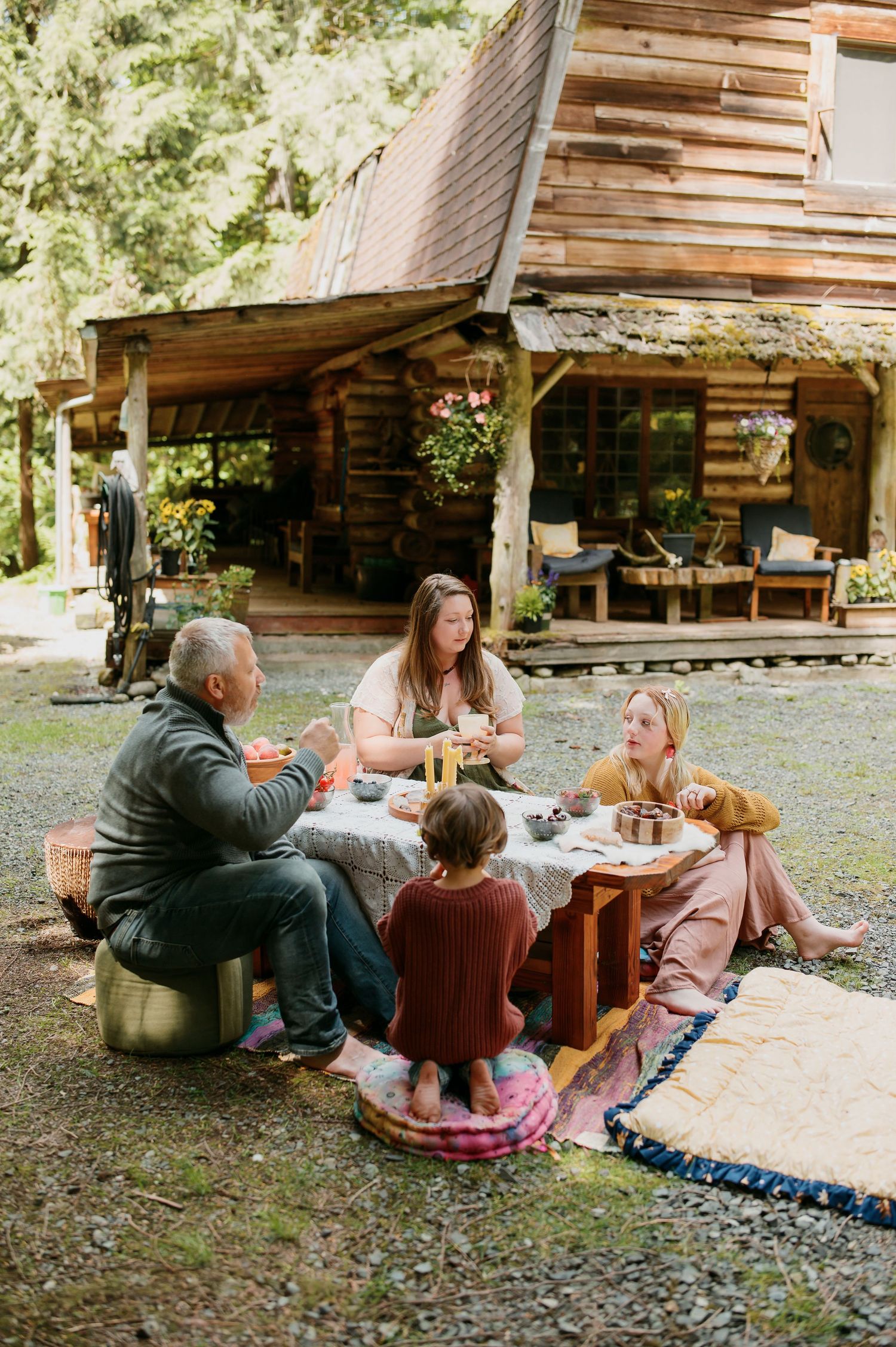 A family enjoys an outdoor meal together on a rustic patio in front of a cozy log cabin in the woods.