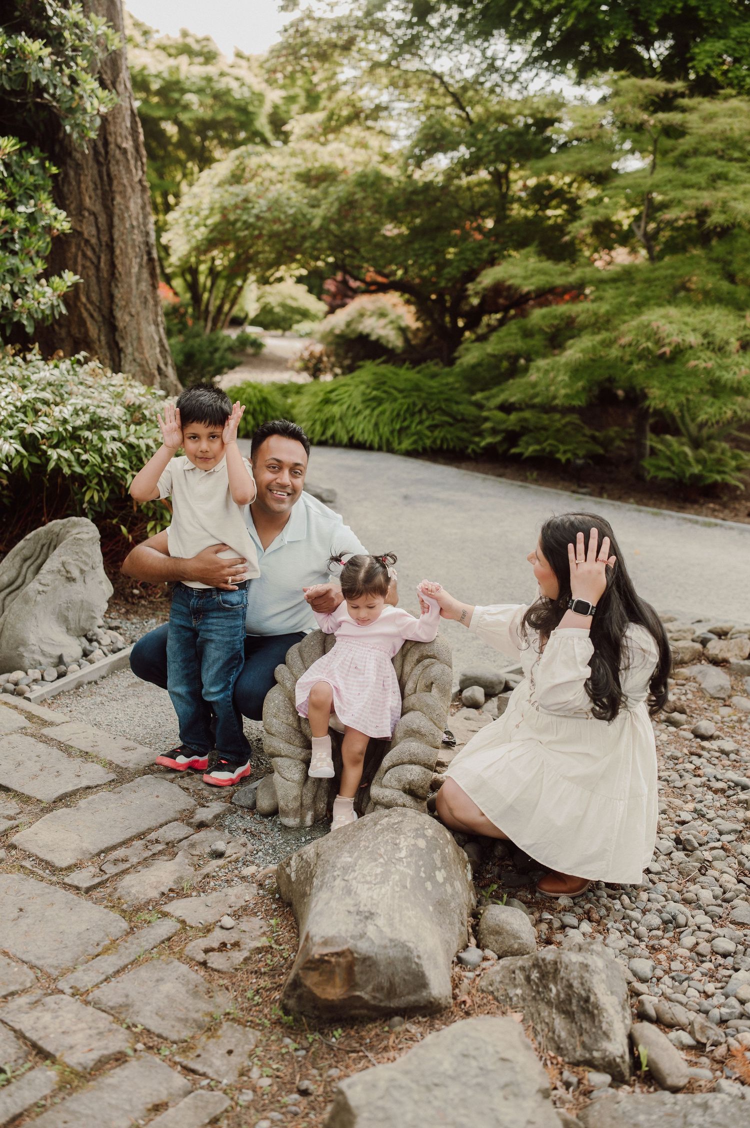 Family of four posing on rocks in a lush Japanese garden, parents waving with two young children.