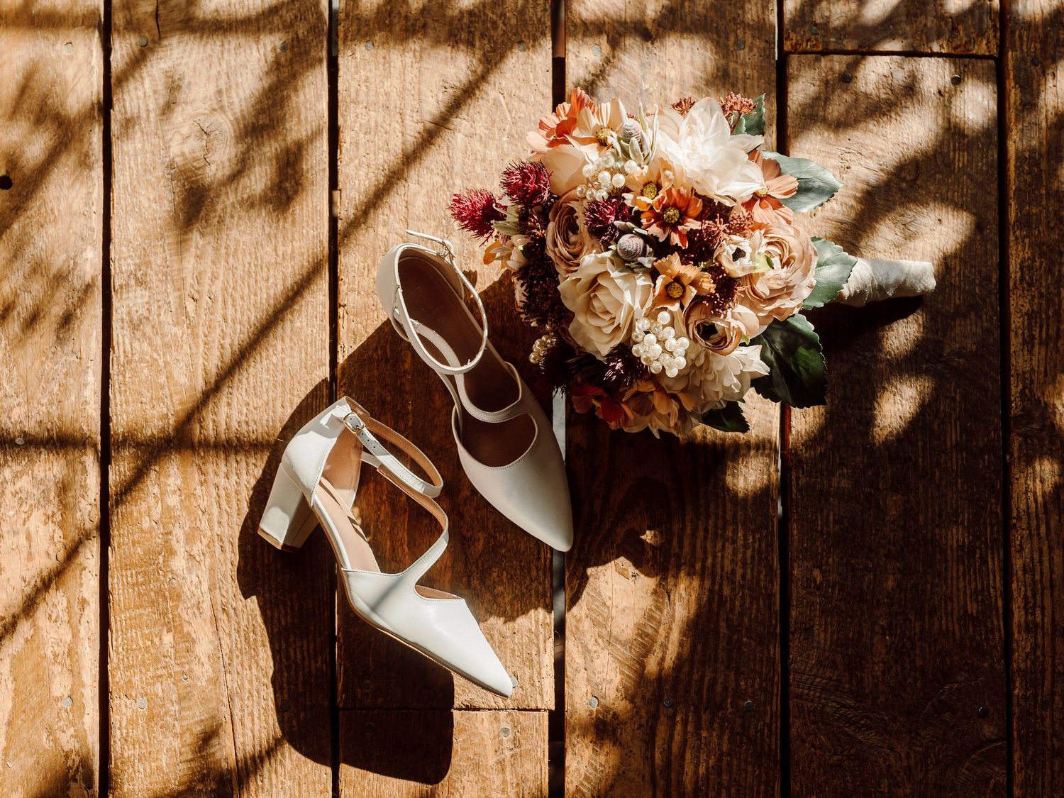 Wedding shoes and a romantic dried flower bouquet in warm sunlight on wooden flooring.