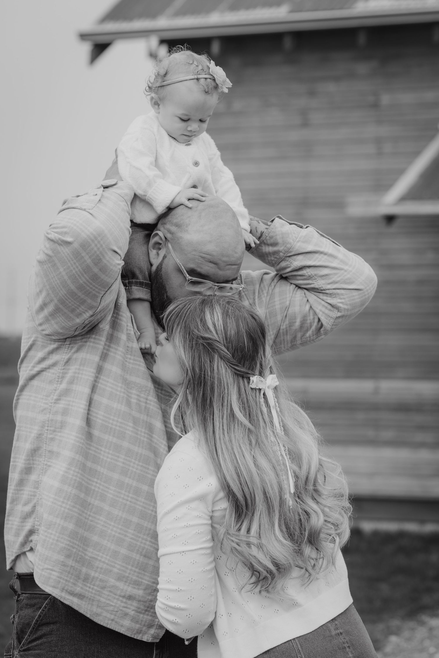 Black and white photo of a man with a baby on his shoulders and a woman leaning in, standing near a barn.