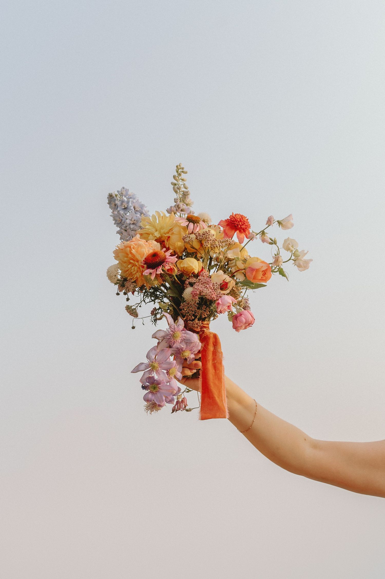 A hand in an orange glove holds a delicate bouquet of pink, orange and yellow flowers against a light beige background.