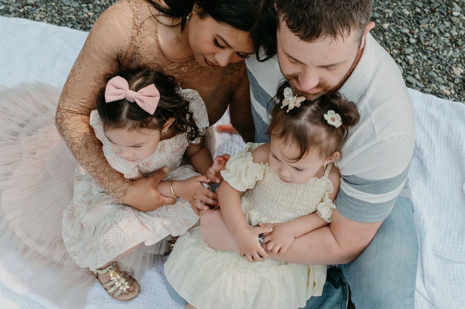 A family sits together on a white blanket outdoors, with two small children wearing matching bows and light dresses.