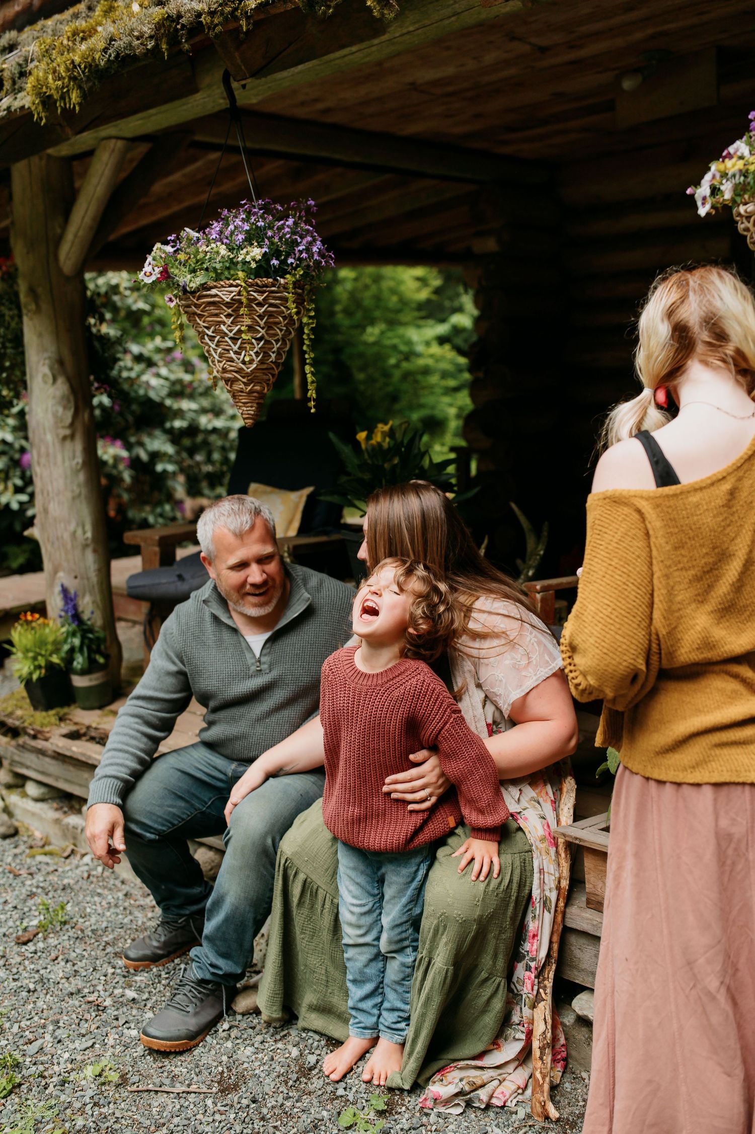 A candid outdoor moment shared between family members on a porch with hanging flowers and rustic wooden beams above.