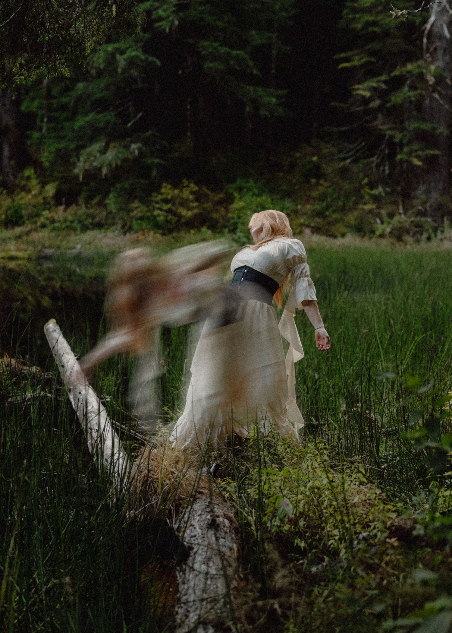 A blonde woman in a white dress with a dark sash walks through a lush green forest meadow, with a blurred figure nearby.