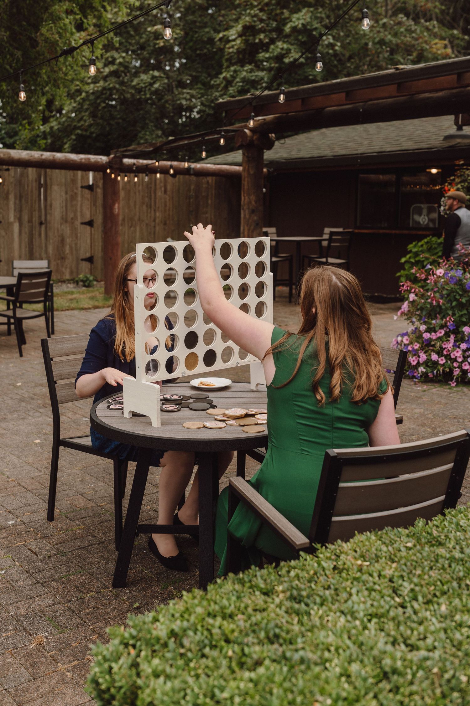 People playing giant Connect Four outdoors in a garden patio setting with string lights and greenery.