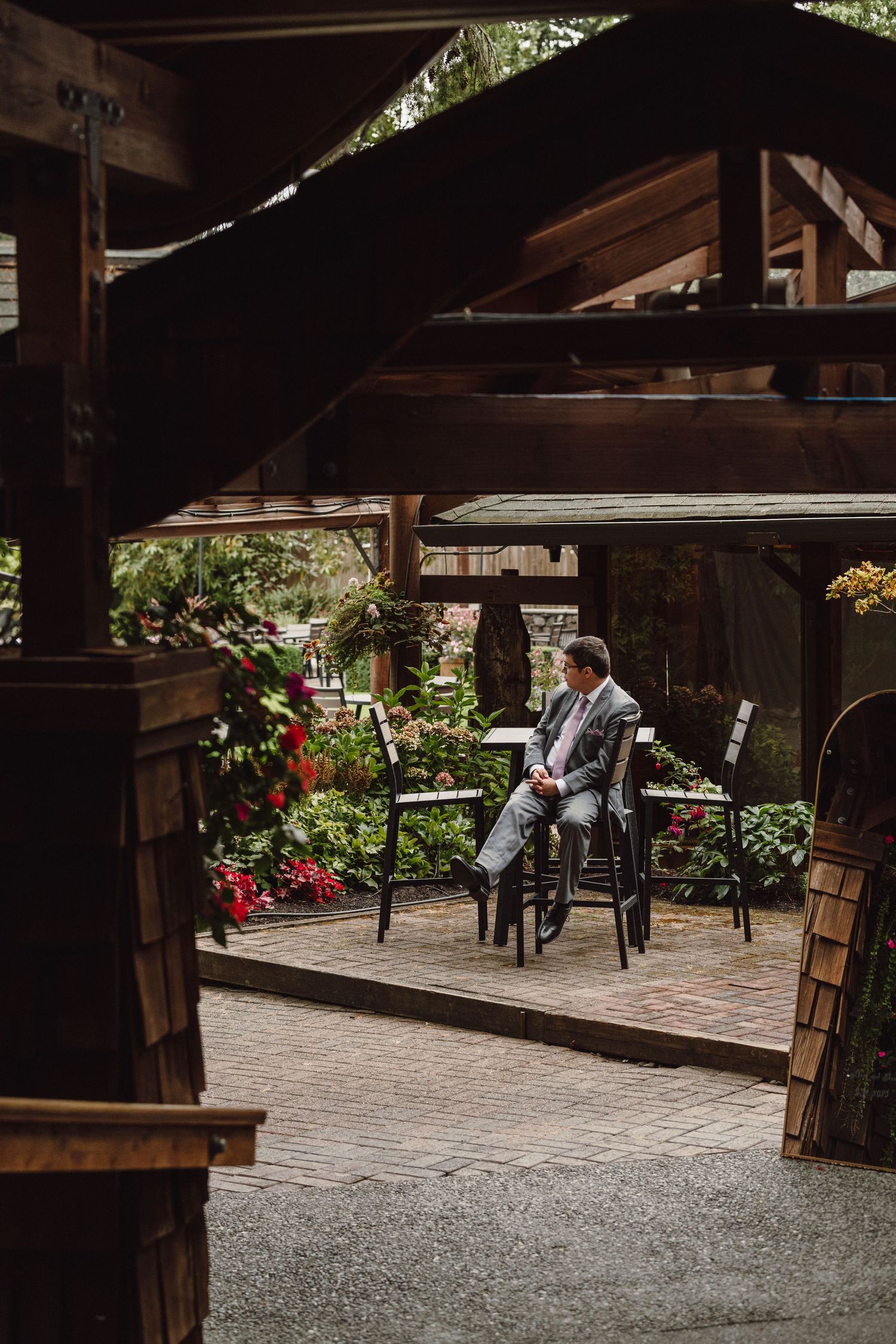 A rustic wooden gazebo with colorful flowers surrounds a figure seated in a chair on a brick patio.