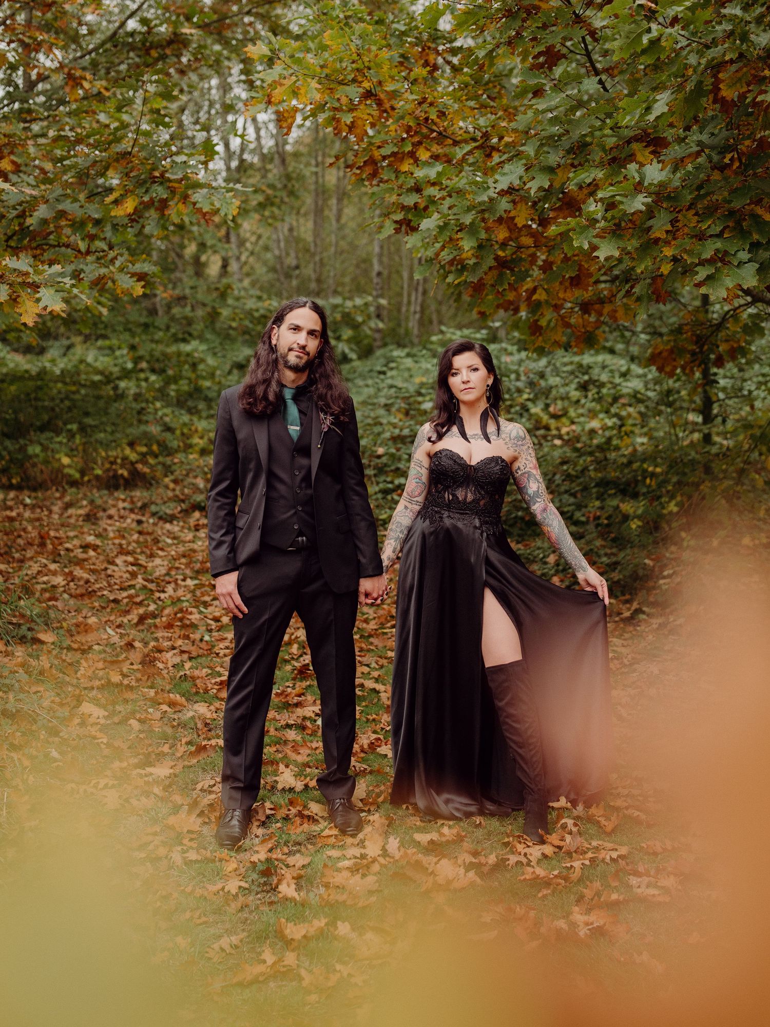 Two people in dark formal attire pose together in a wooded autumn setting with fallen leaves on the ground.