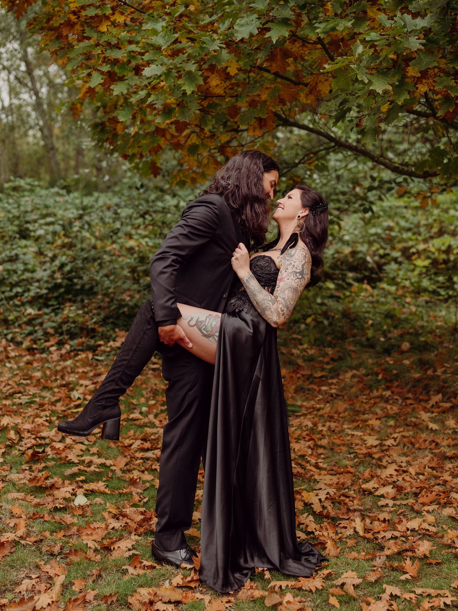 A romantic dip kiss between two people in dark formal wear surrounded by fall foliage.