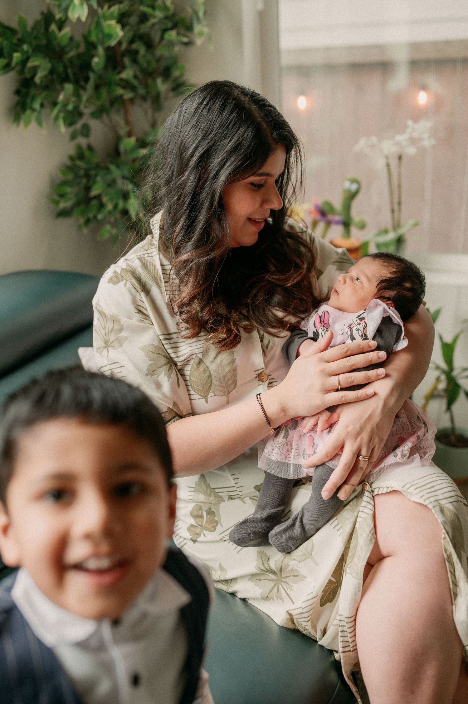 A tender family moment captured indoors with a baby and young child in a cozy living room setting with plants and soft lighting.