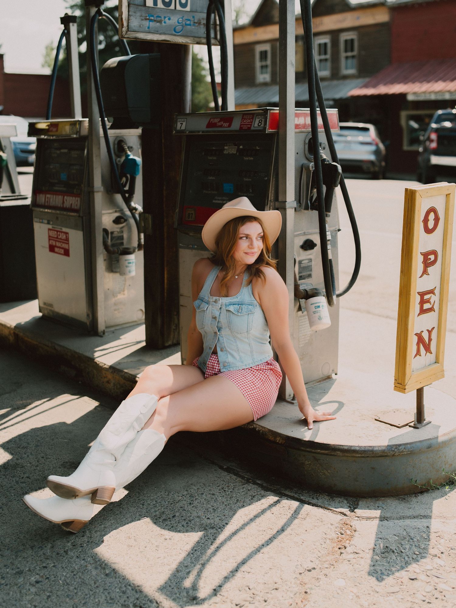 A woman in a summer dress and hat sits casually on a gas station pump island in warm sunlight.