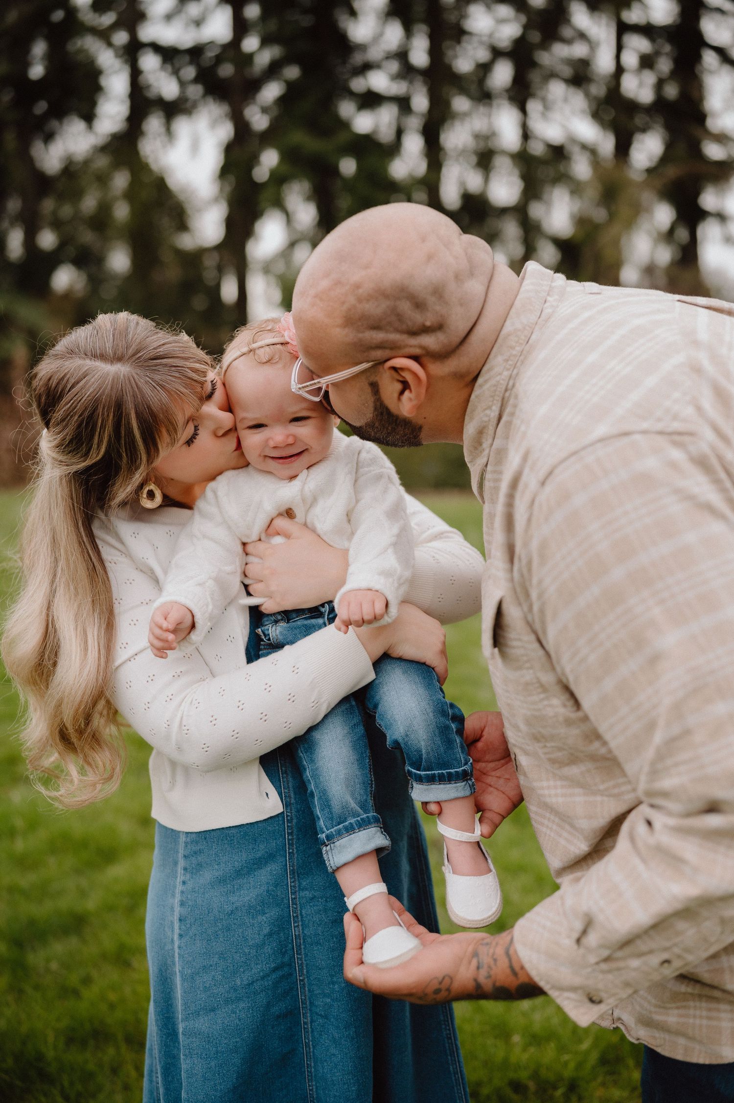 A couple kisses their smiling baby girl outdoors, with the mother in a white sweater and blue skirt and father in a plaid jacket.