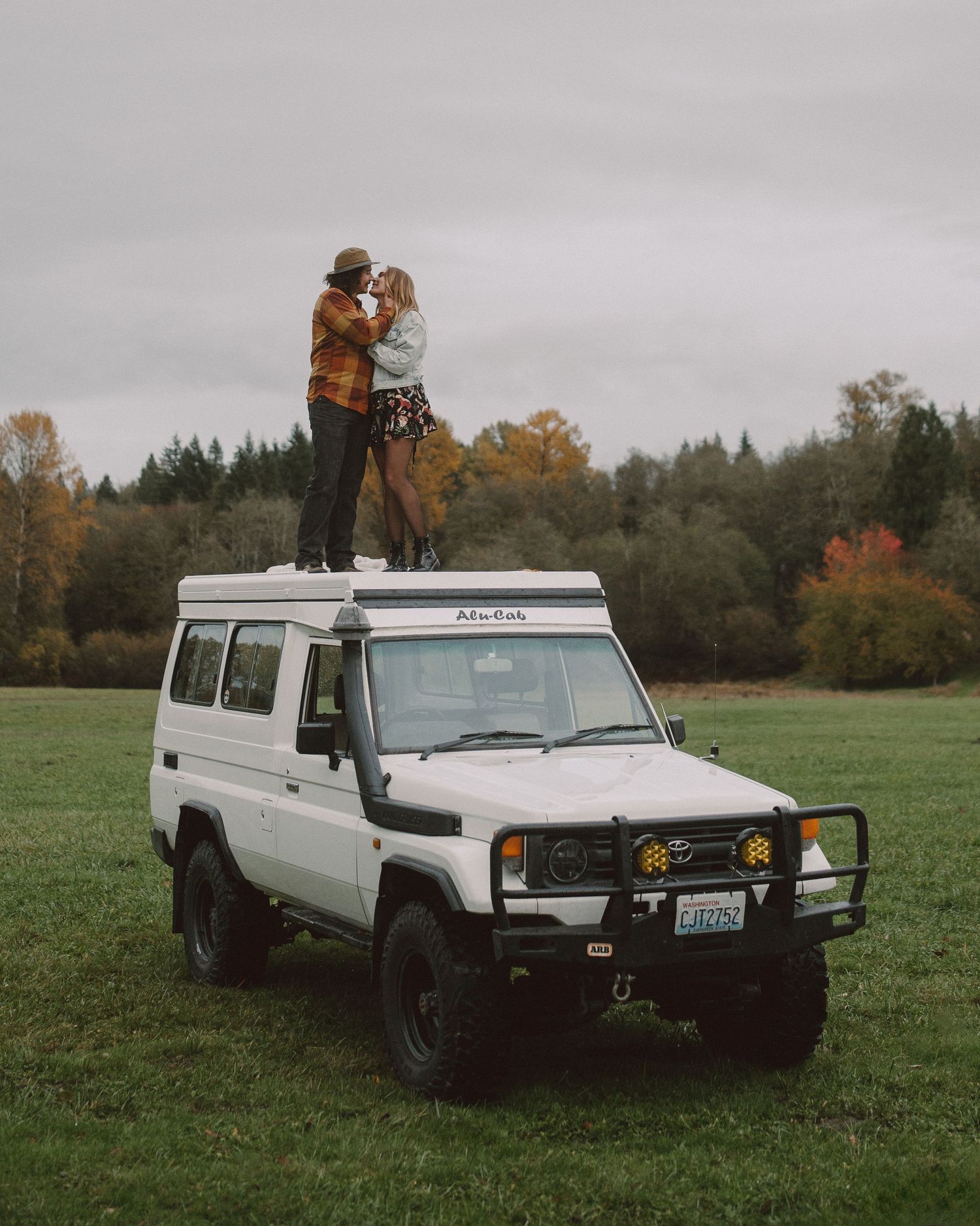 A couple stands atop a white Toyota Land Cruiser in a fall field.