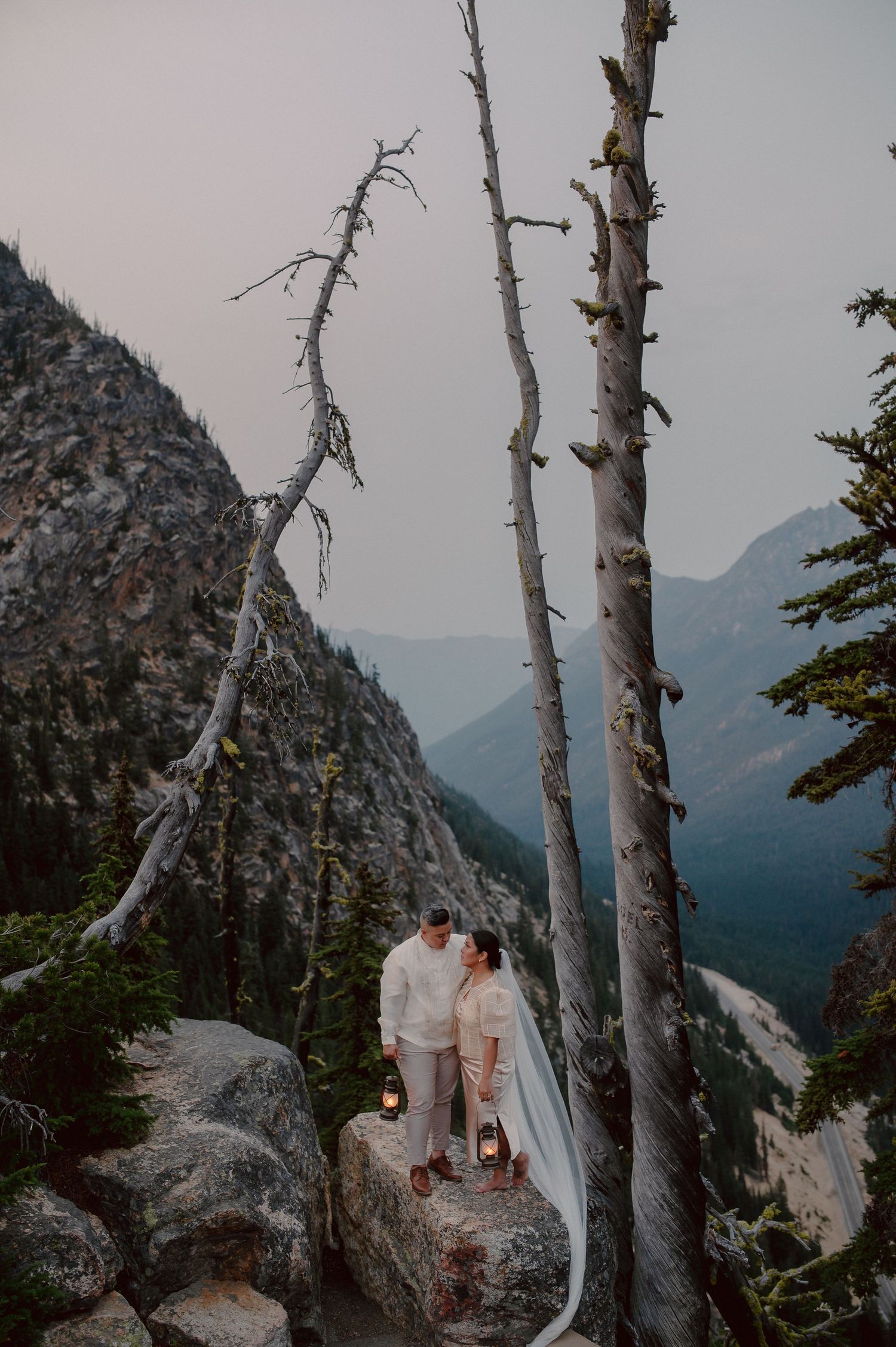 Two figures stand on a rocky mountain ledge surrounded by tall birch trees and misty mountain views at dusk.