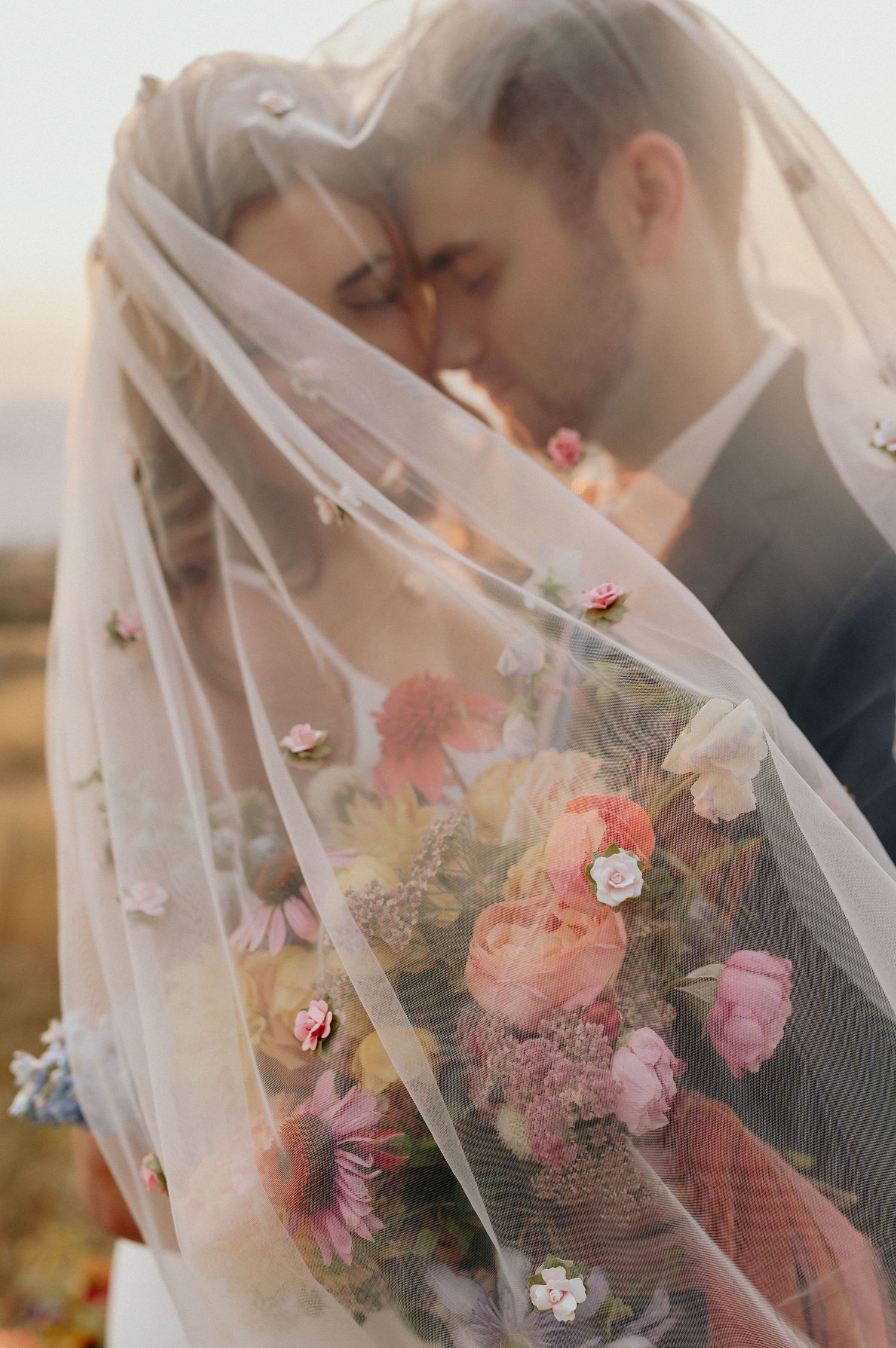 A romantic wedding moment captured through delicate white veil with pink and white floral bouquet in soft focus.