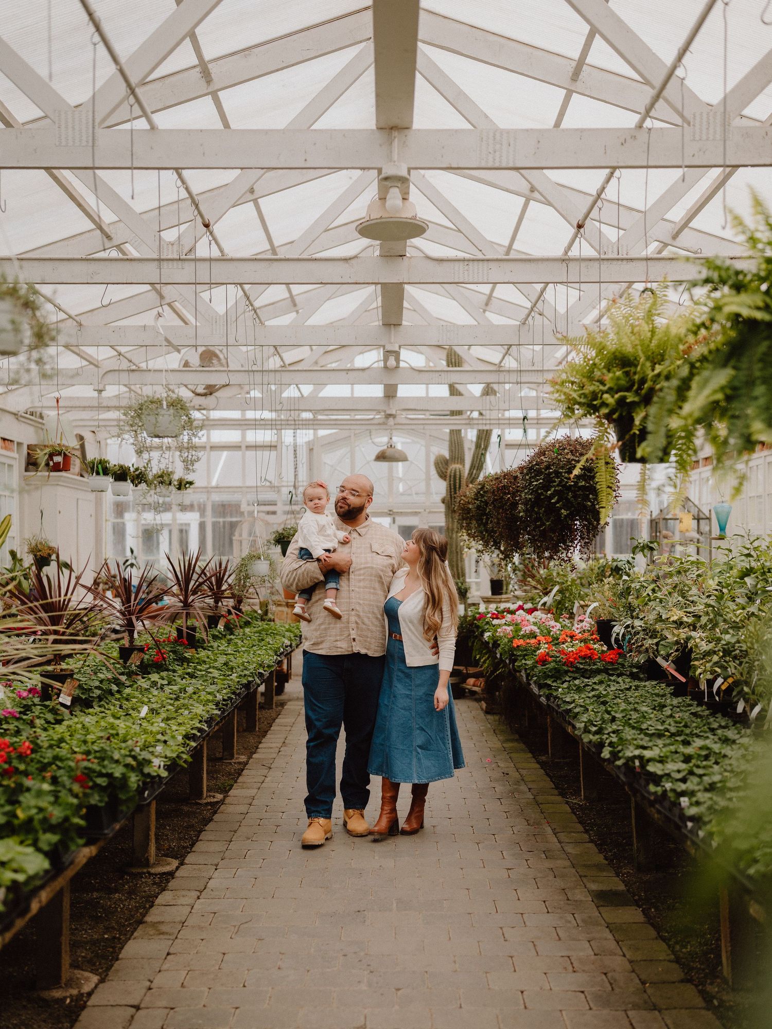 A family with a baby strolls through a lush greenhouse filled with plants and flowers.