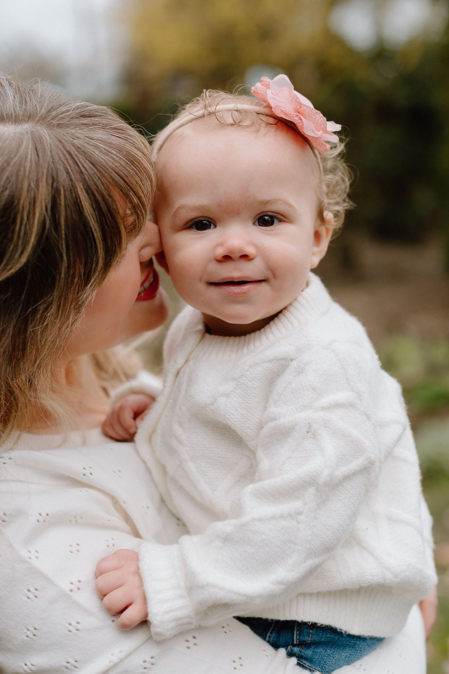 A mother in a white sweater holds her smiling baby girl wearing a white sweater and pink bow headband outdoors.