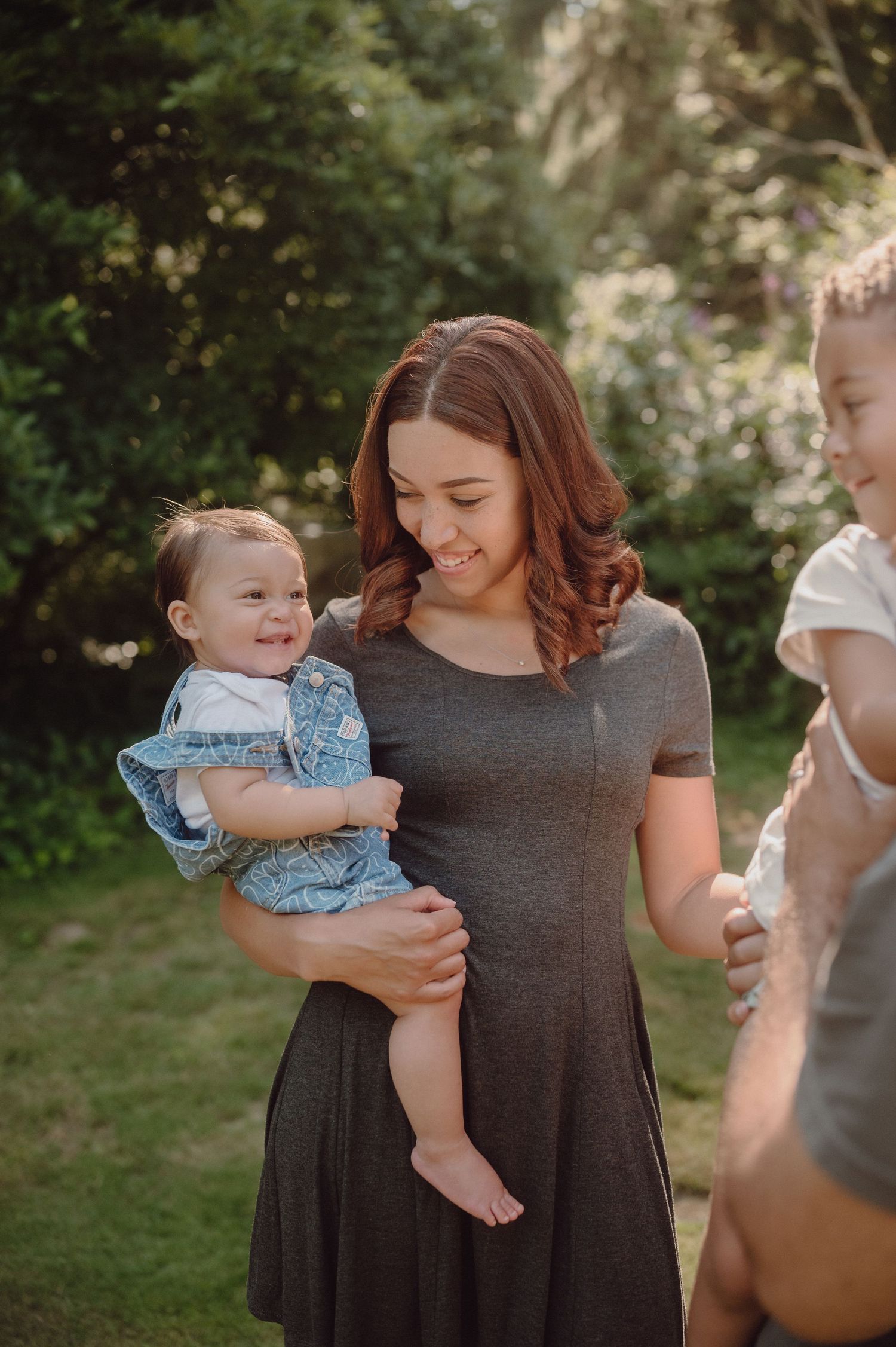 A smiling woman in a gray dress holds a happy baby in a denim outfit outdoors in a lush green garden.