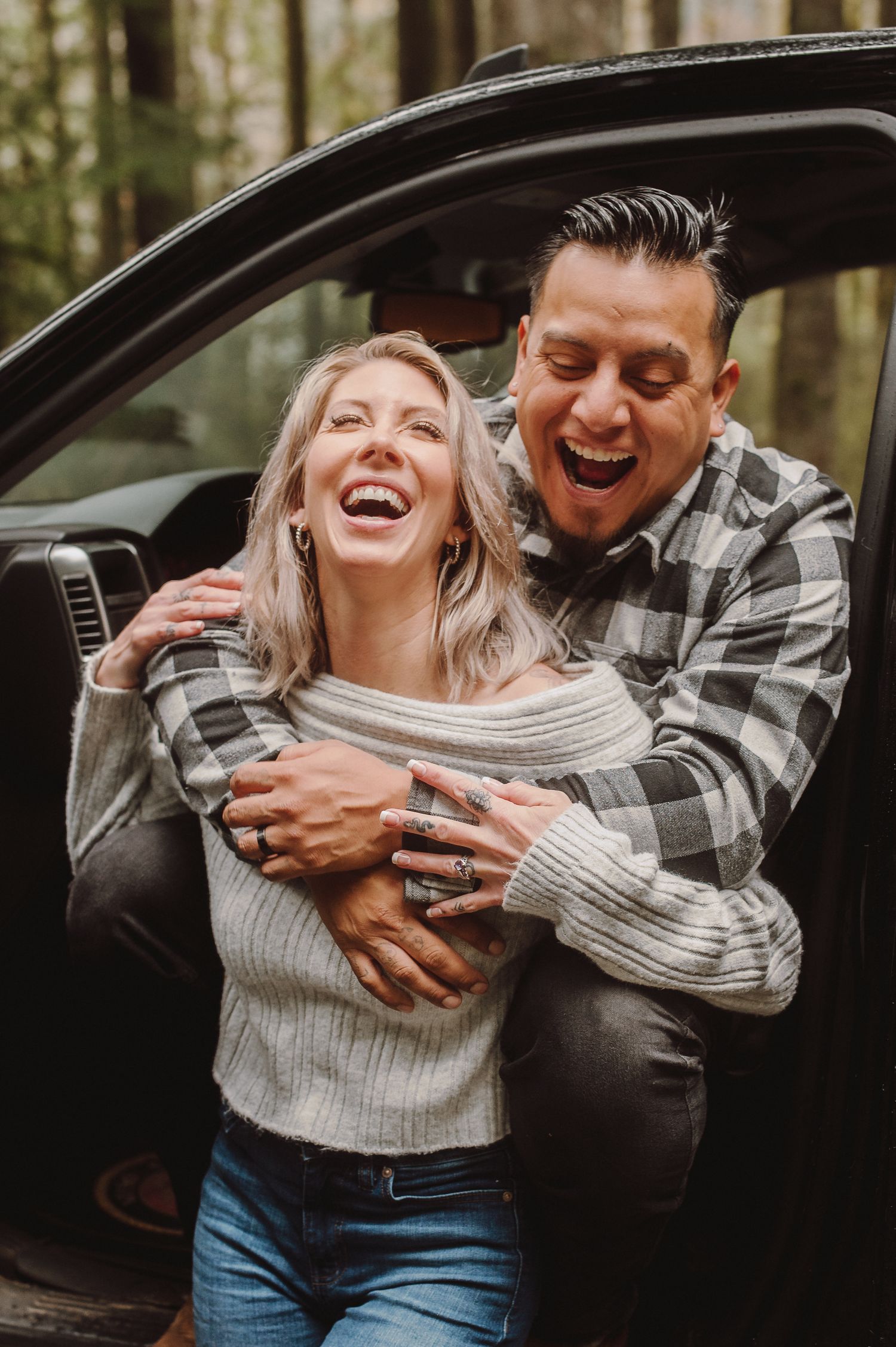 A joyful couple shares a candid moment of laughter together while standing by their car in a wooded setting.