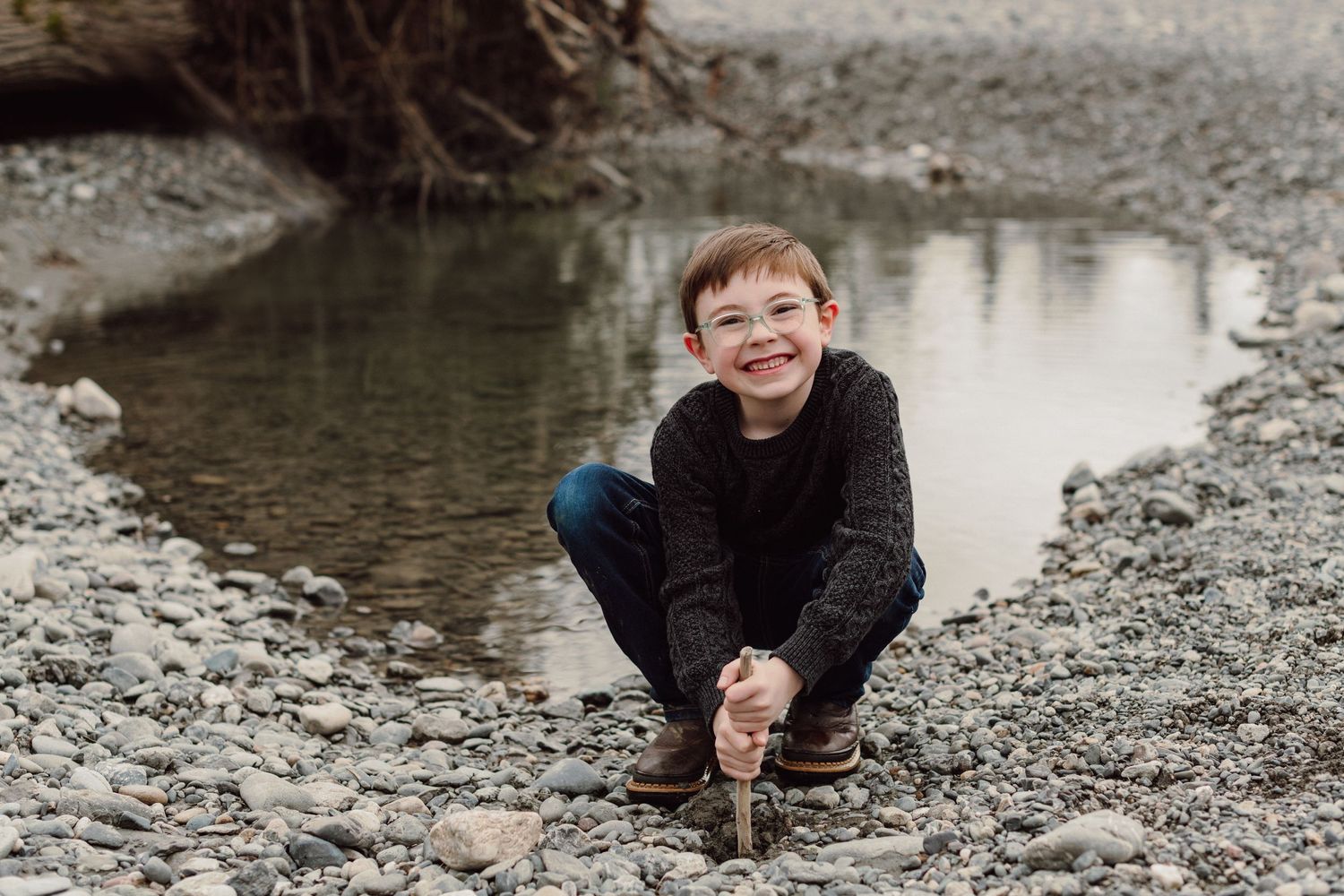 A smiling child crouching by a small pond or stream surrounded by rocks.