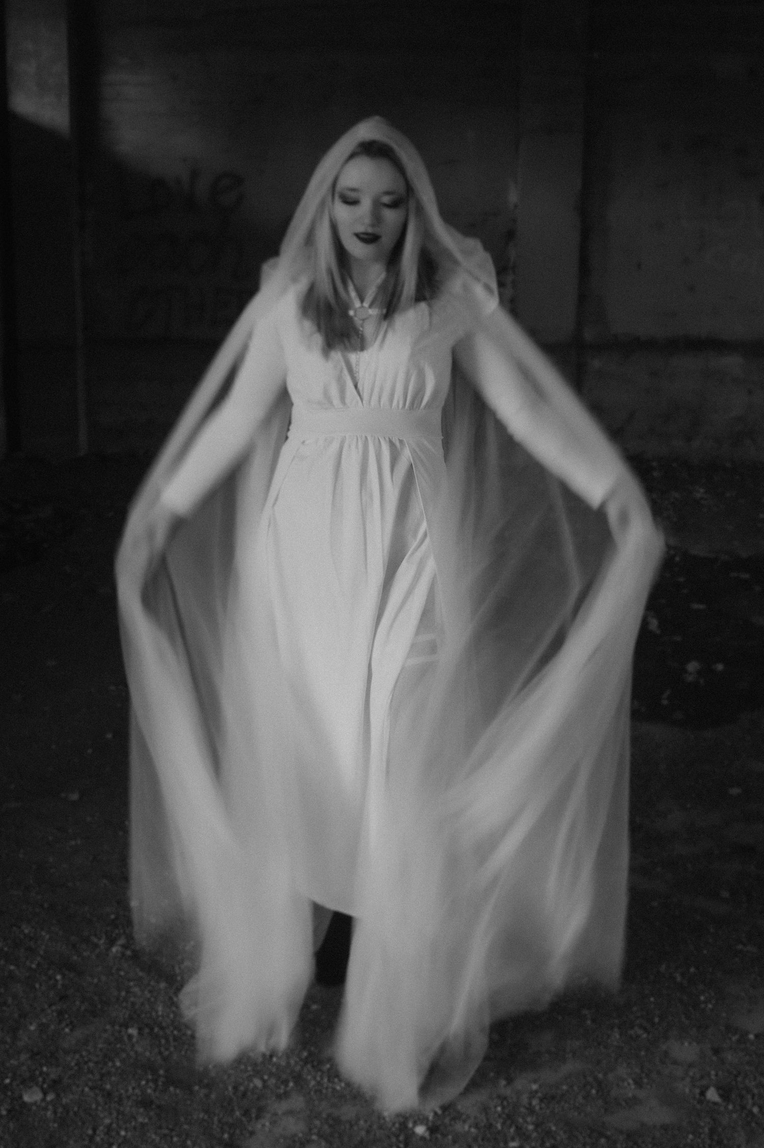 A ghostly figure in a flowing white dress stands dramatically in a dark setting in this black and white photograph.