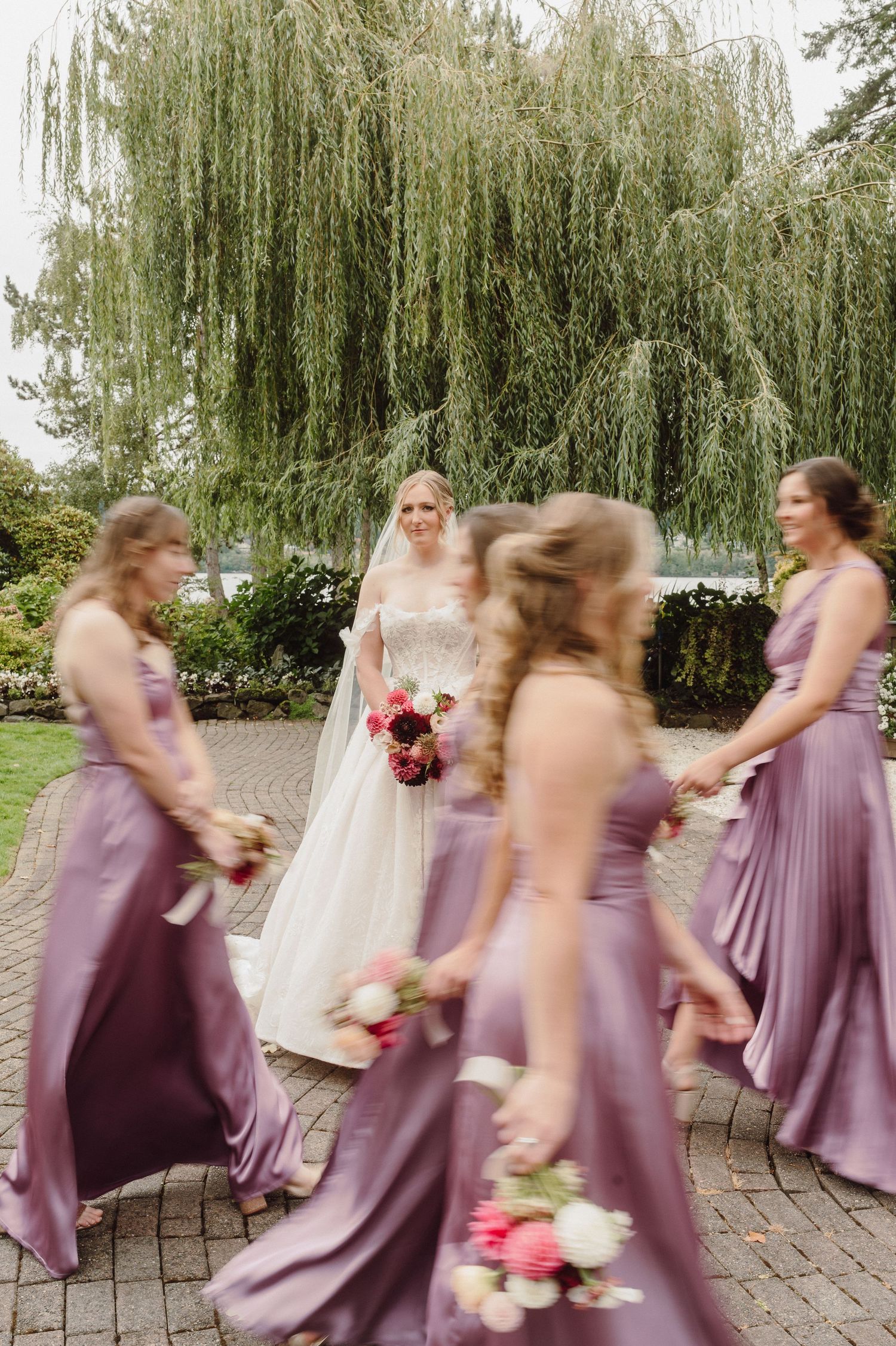 Bridesmaids in flowing lilac dresses create motion blur while walking in sequence under a weeping willow tree.