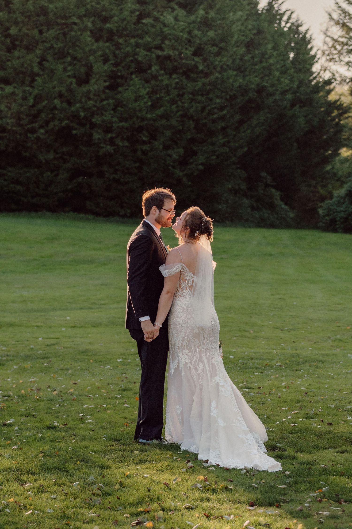 A wedding couple stands together on a lush green lawn at sunset.