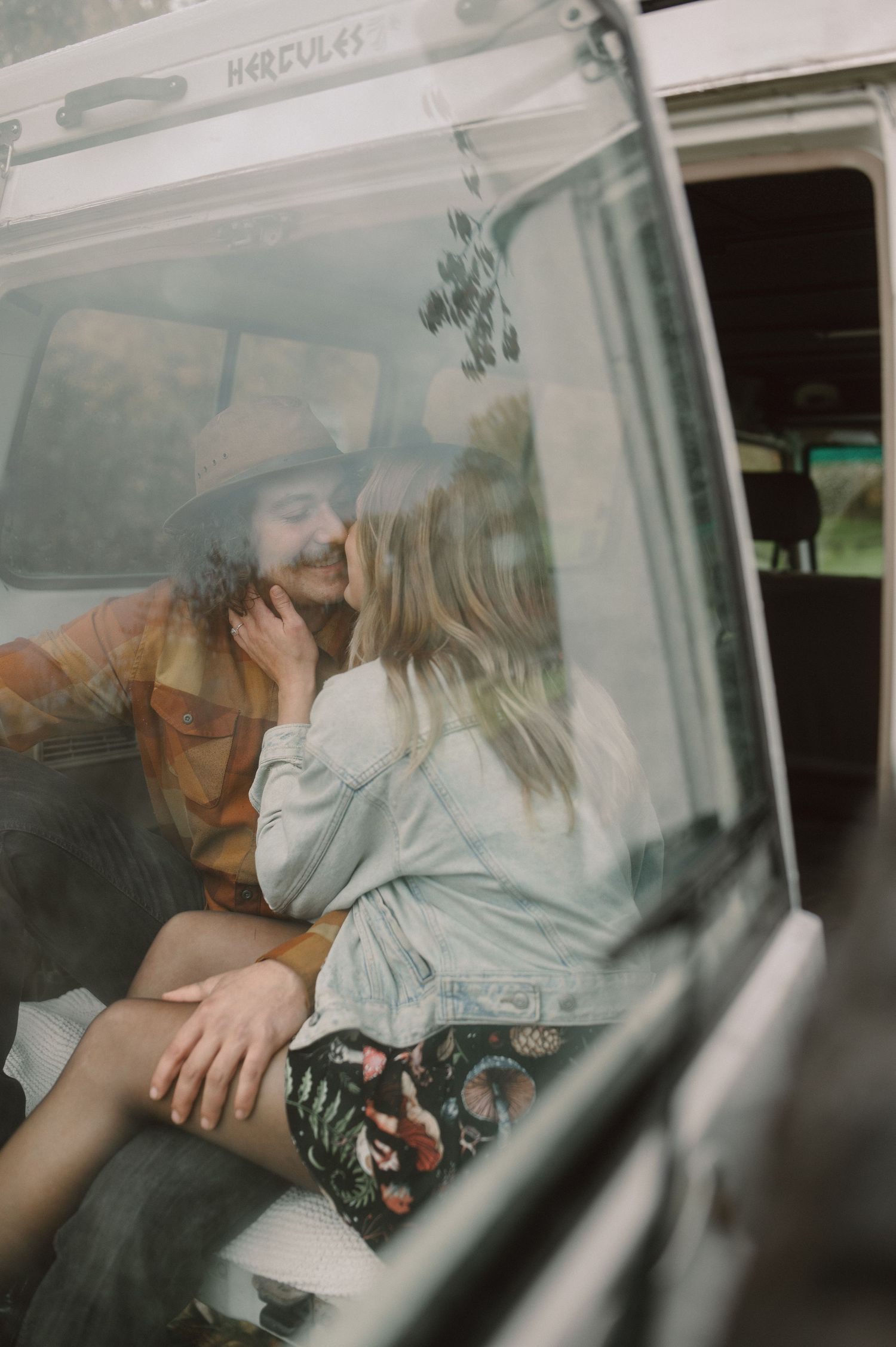 A couple share an intimate moment in the backseat of a vintage van, shot through the window with a dreamy film aesthetic.