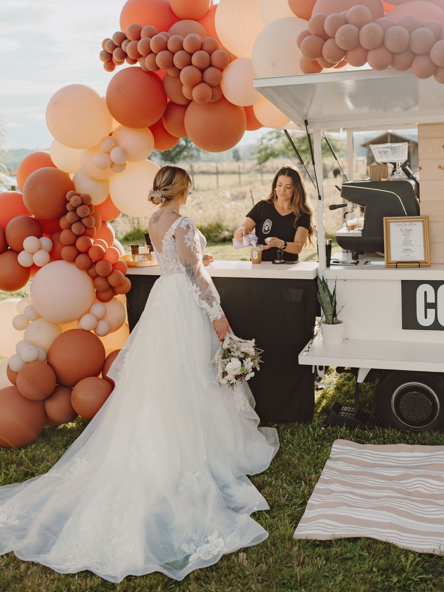 Bride in white lace gown stands at an outdoor coffee cart adorned with terracotta and blush balloon arch at wedding.
