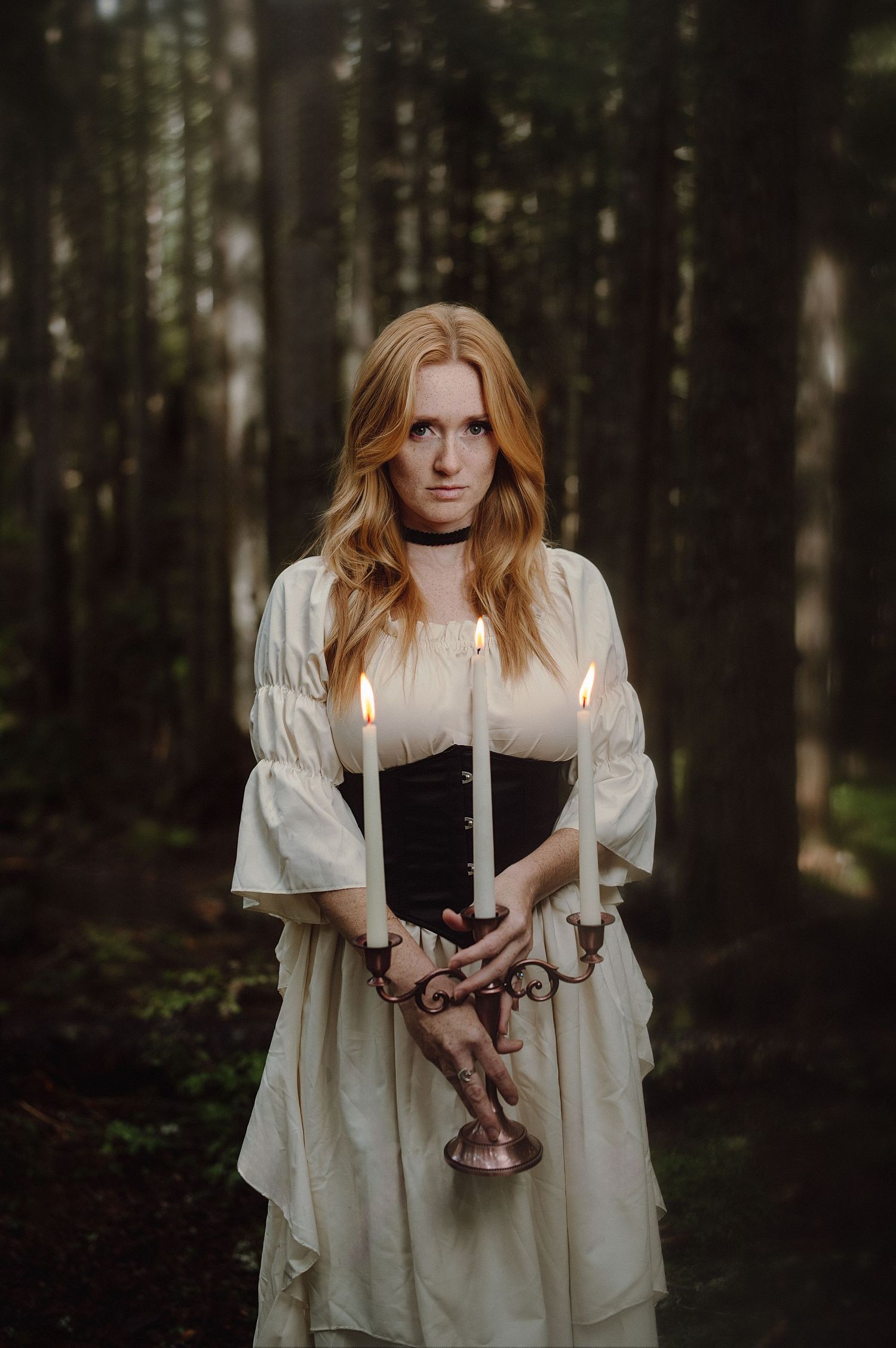 A figure in a white vintage dress holds lit candles in a dark moody forest setting creating an ethereal atmosphere.