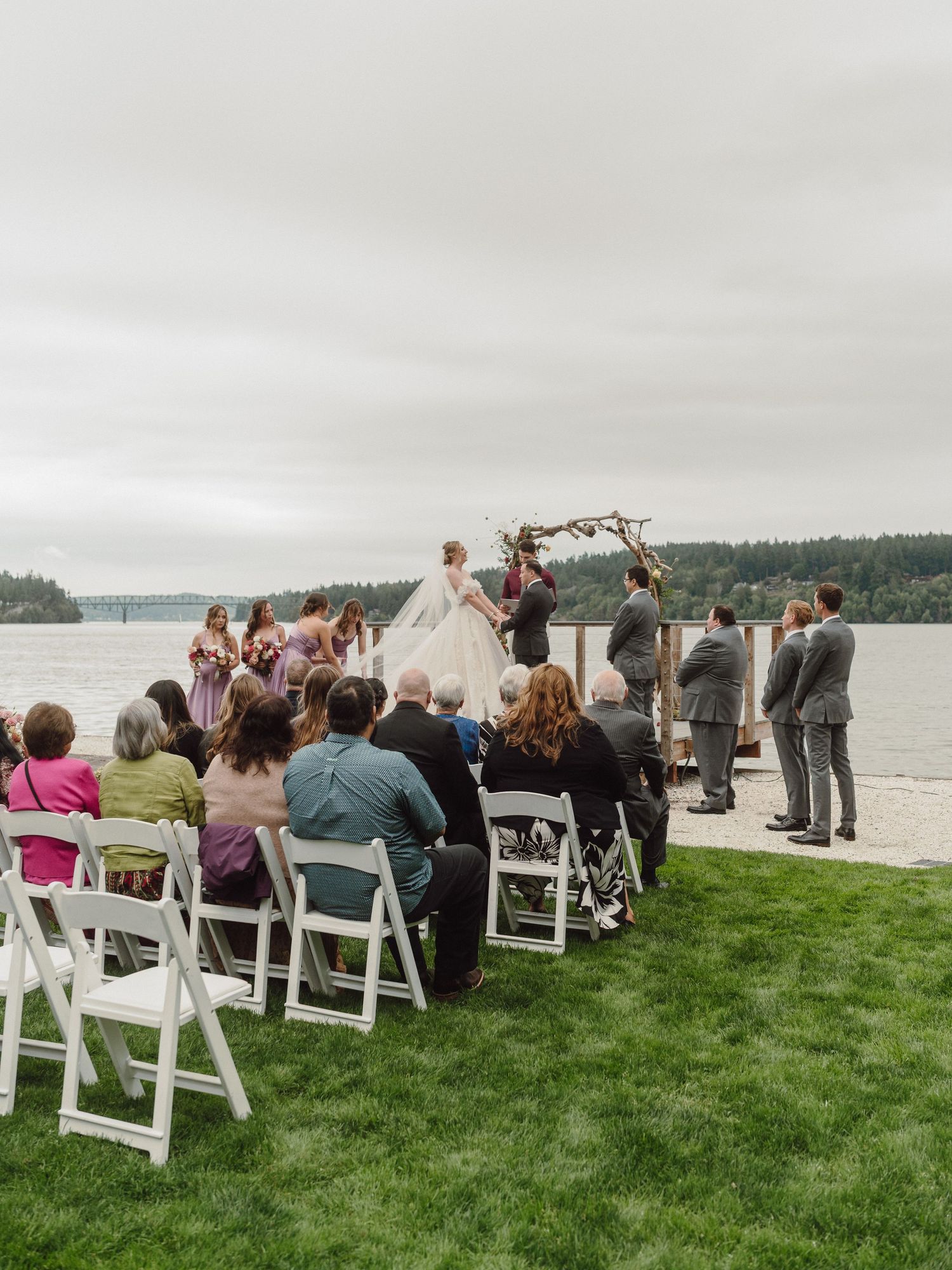 Guests seated in white chairs on green grass overlooking a waterfront during an outdoor wedding ceremony.