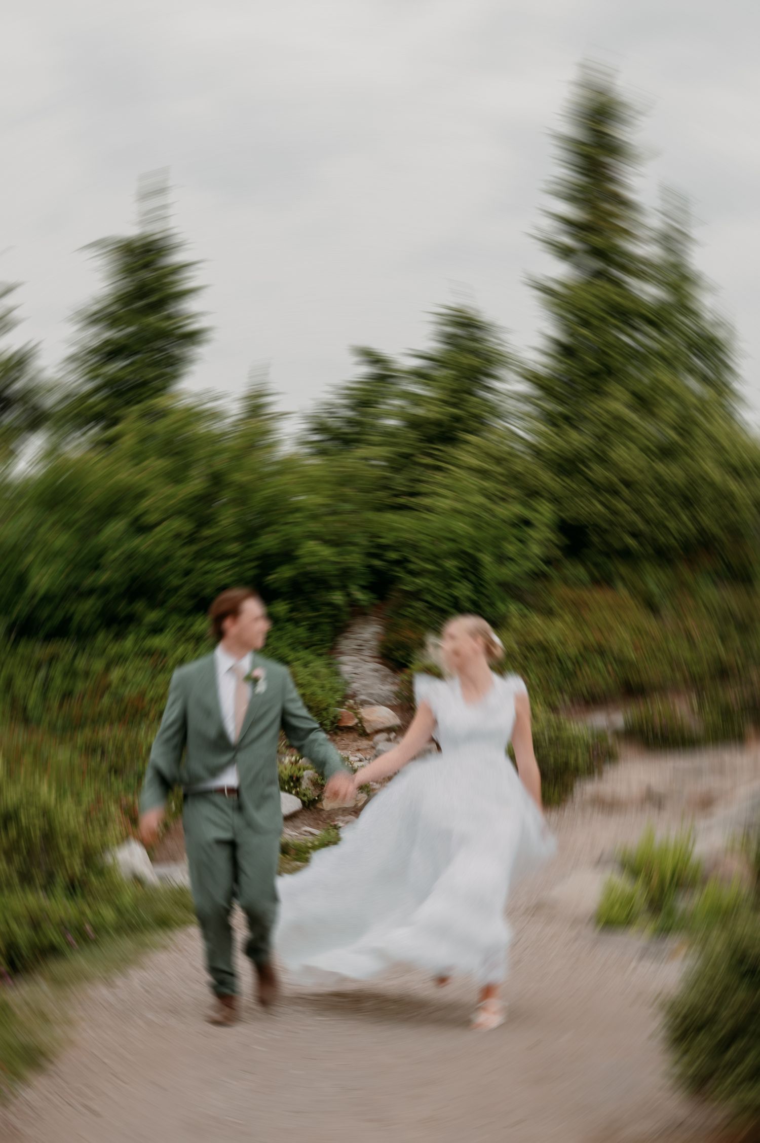 A couple runs down a path surrounded by evergreen trees during their wedding celebration in a natural setting.