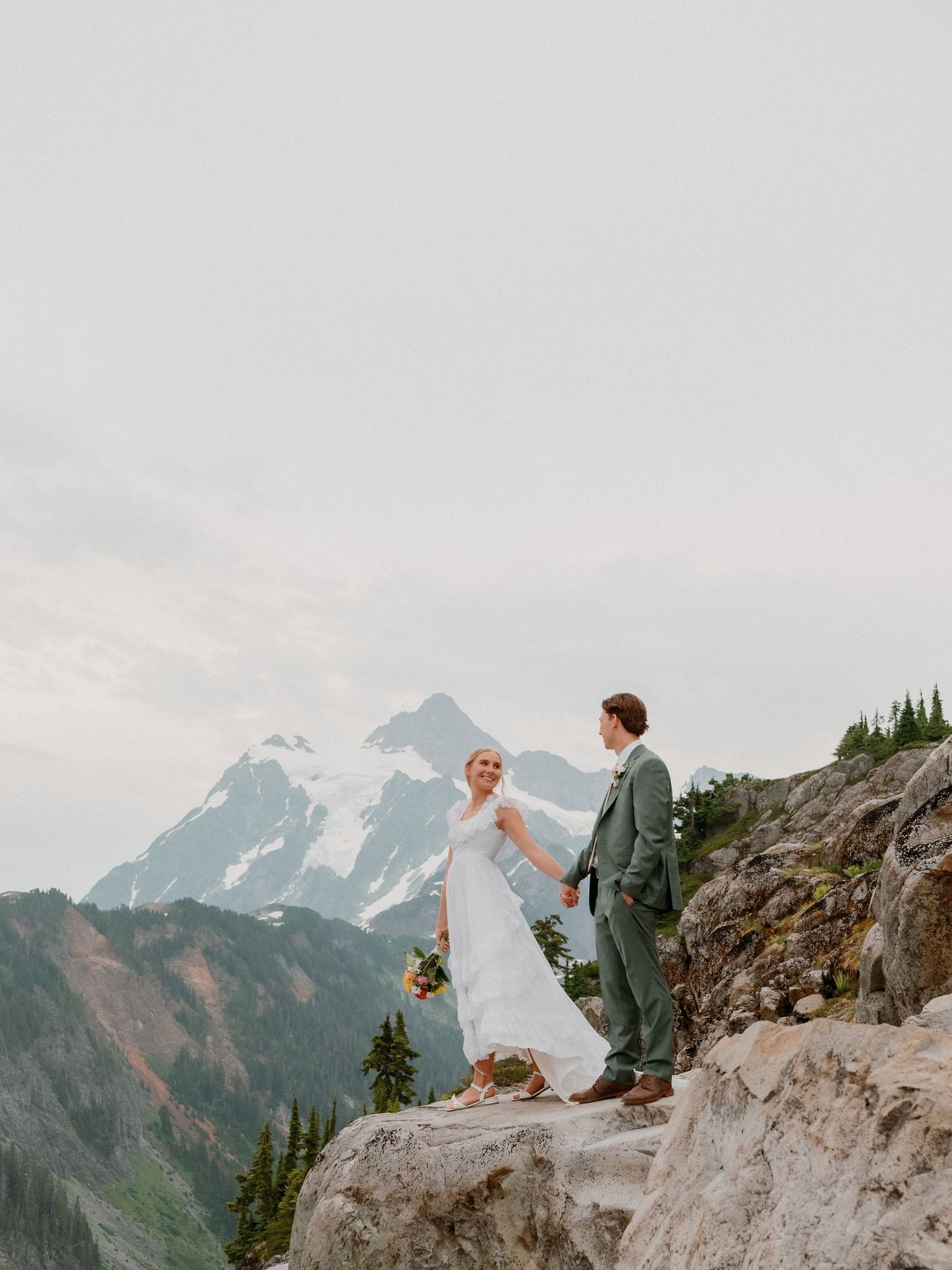A couple in wedding attire stands on a rocky mountain peak with dramatic snow-capped peaks in the background.