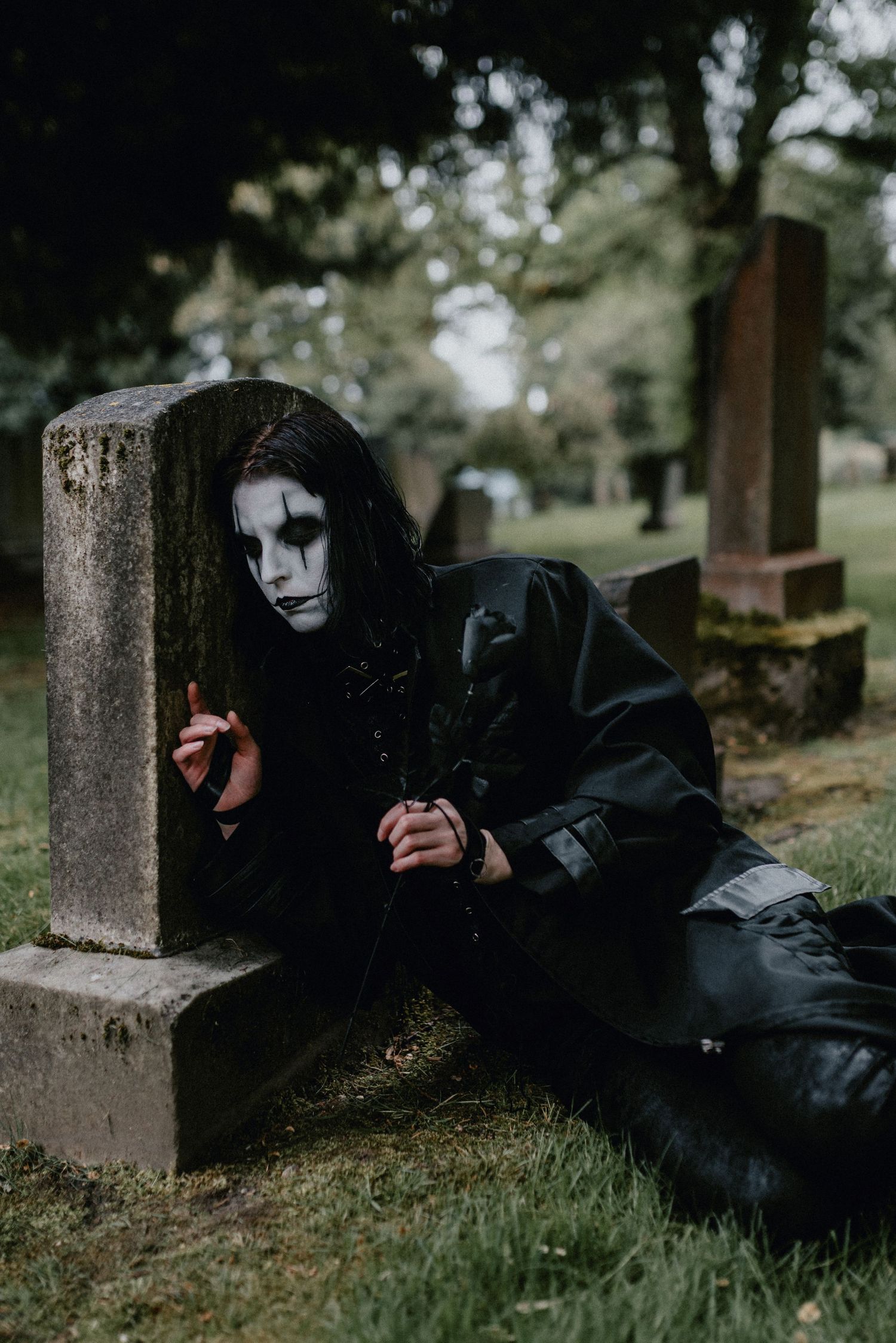 Person in dark clothing and white mask lies on grass near stone pillars in a moody outdoor setting.