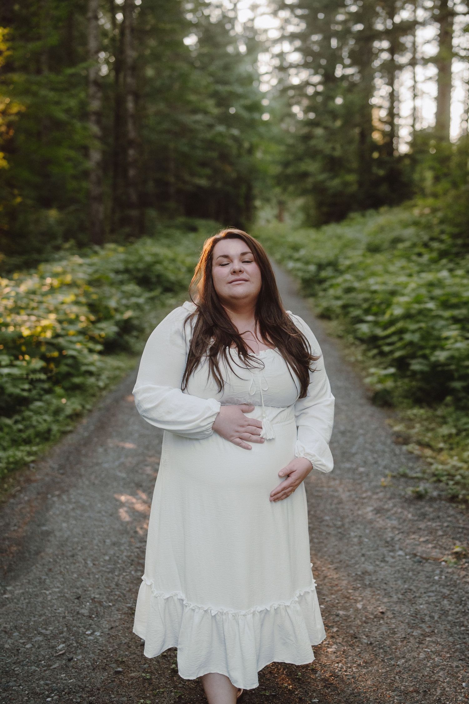 A person in a flowing white dress stands on a gravel path surrounded by lush green forest scenery.