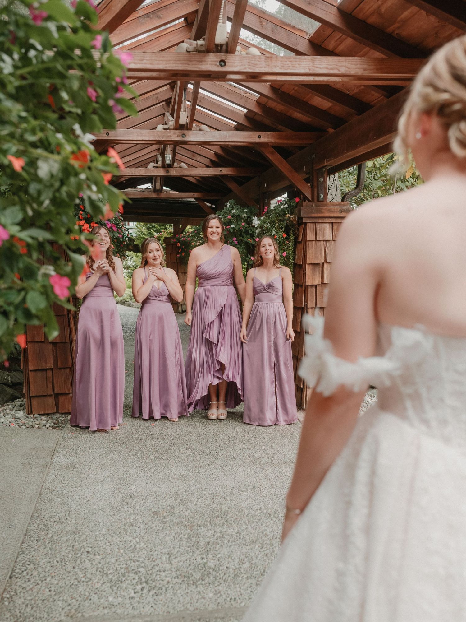 Bridesmaids in flowing mauve dresses share a candid moment under a rustic wooden pergola during wedding celebrations.