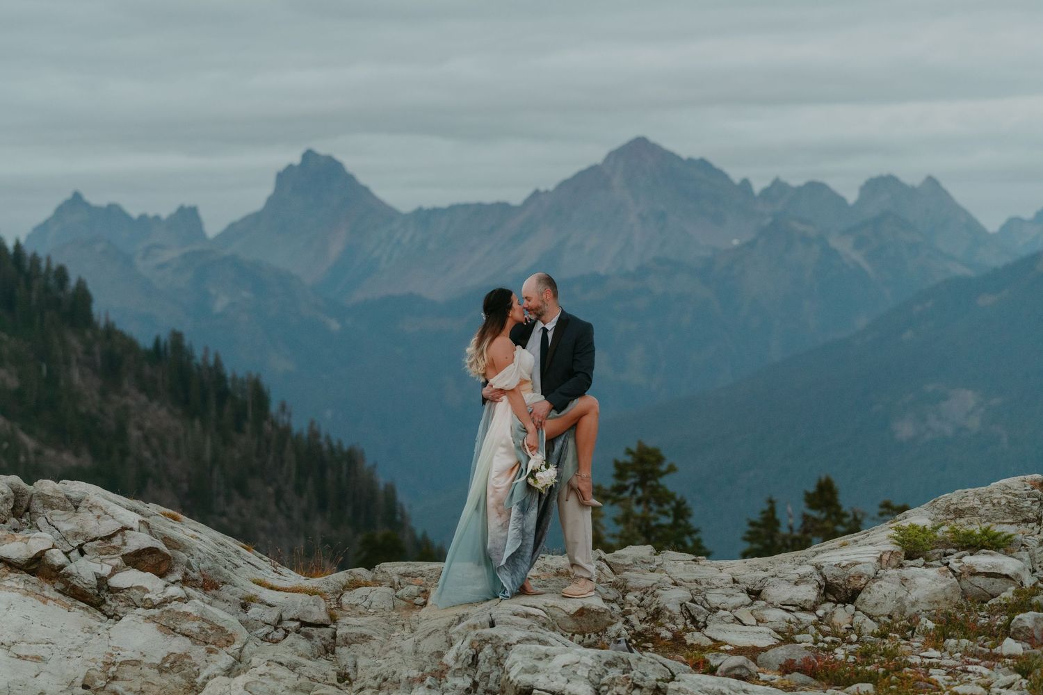 Couple sharing a romantic kiss on rocky mountain summit with dramatic peaks and cloudy sky backdrop.