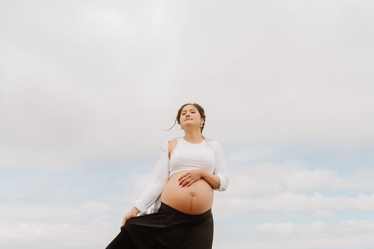 Artistic maternity photo against cloudy sky showing pregnant belly in white top.
