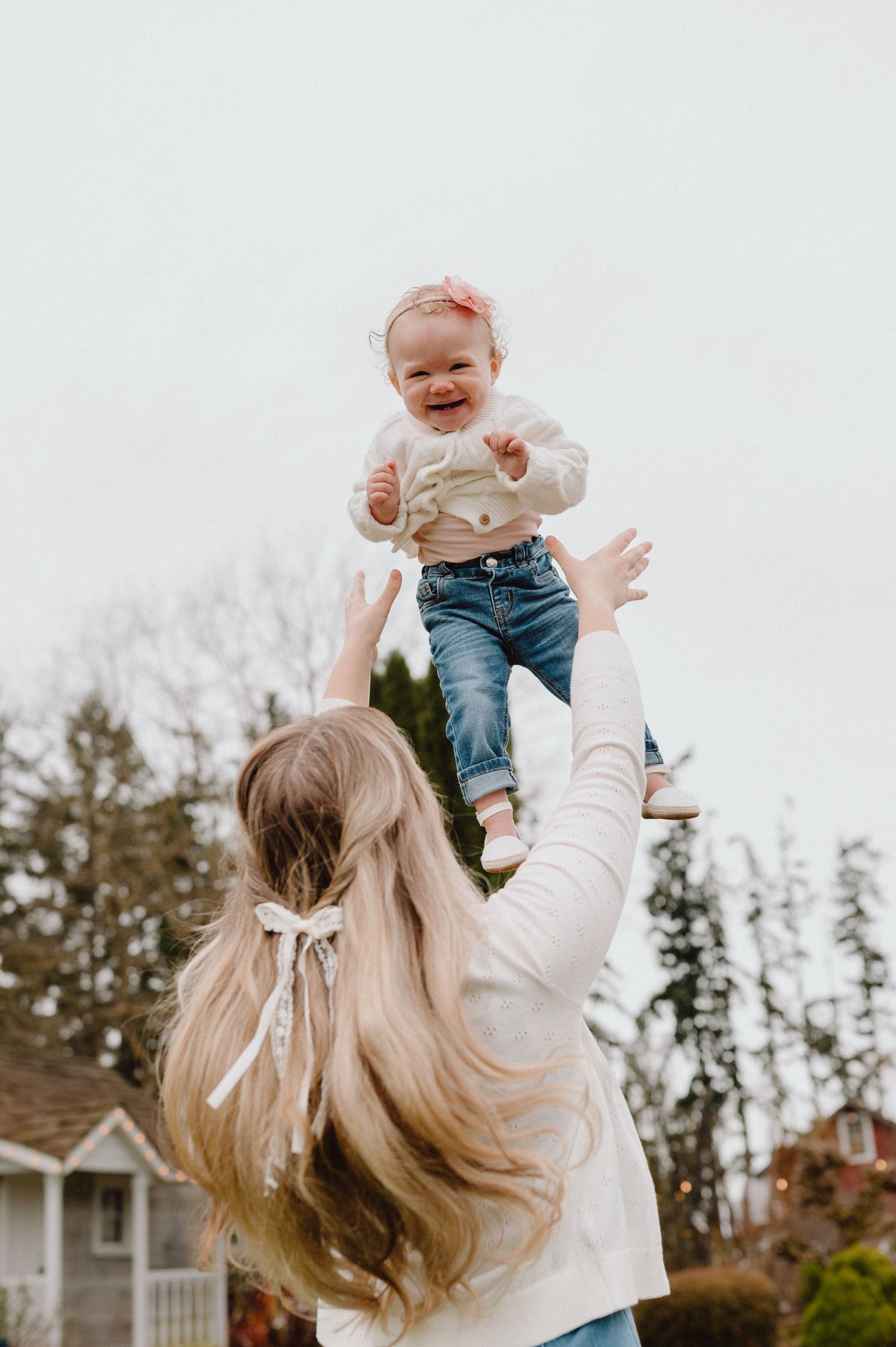 A smiling baby in jeans and white top being tossed in the air by a blonde woman with a bow in her hair outdoors.