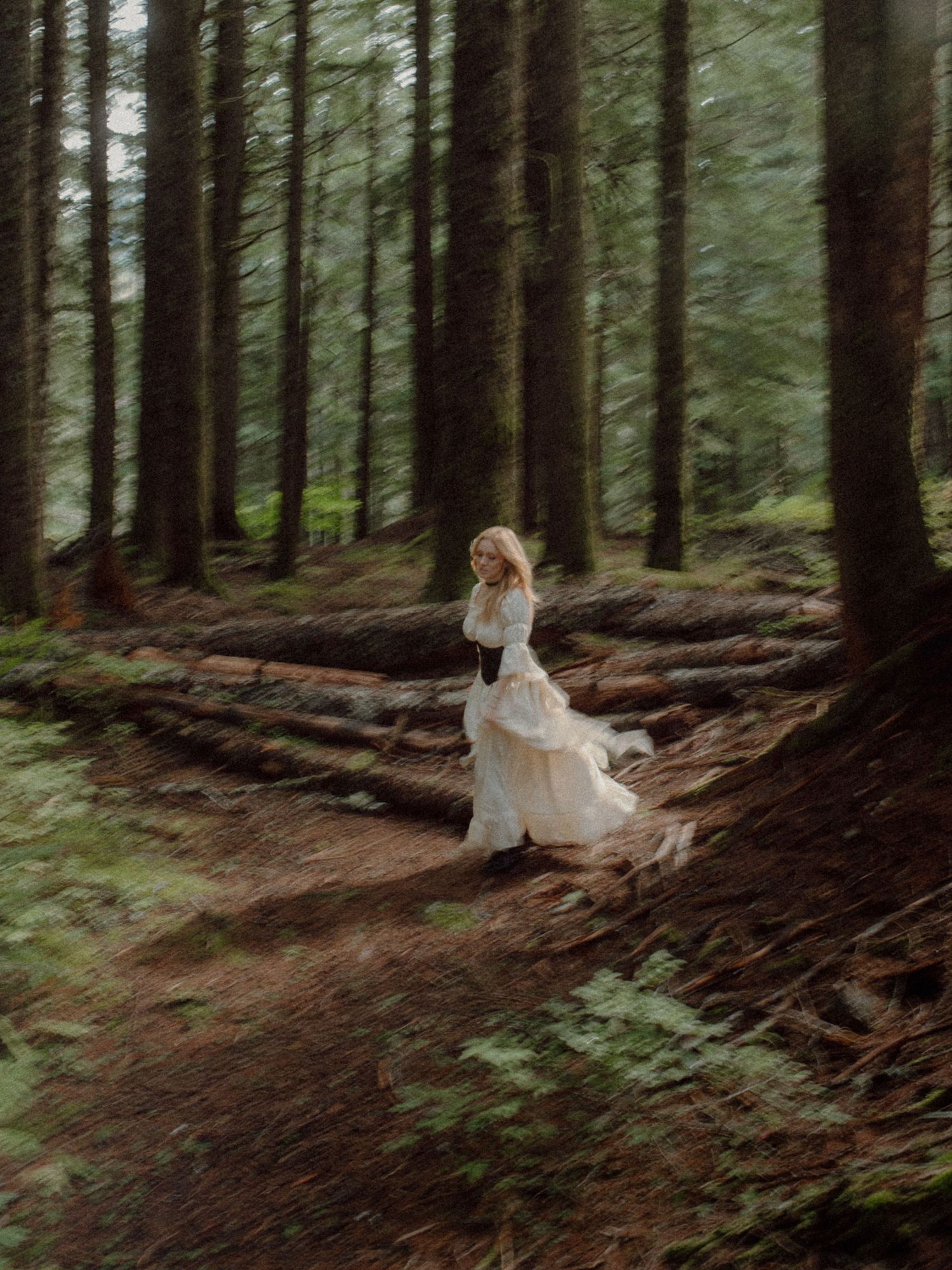 A blonde woman in a flowing white dress walks through a misty forest trail surrounded by tall pine trees and fallen logs.