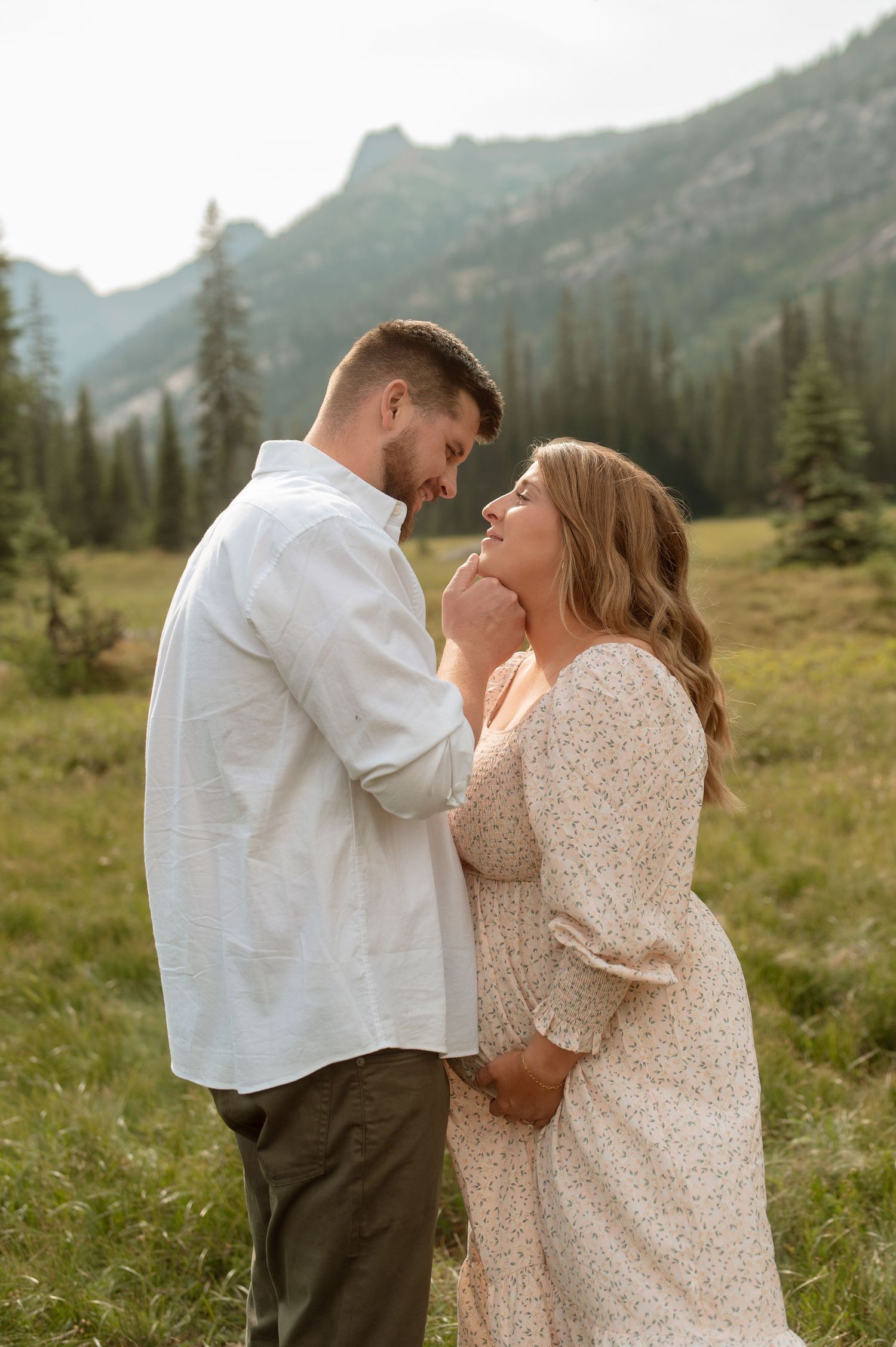 A couple shares an intimate moment in a mountain meadow surrounded by evergreen trees and majestic peaks at sunset.