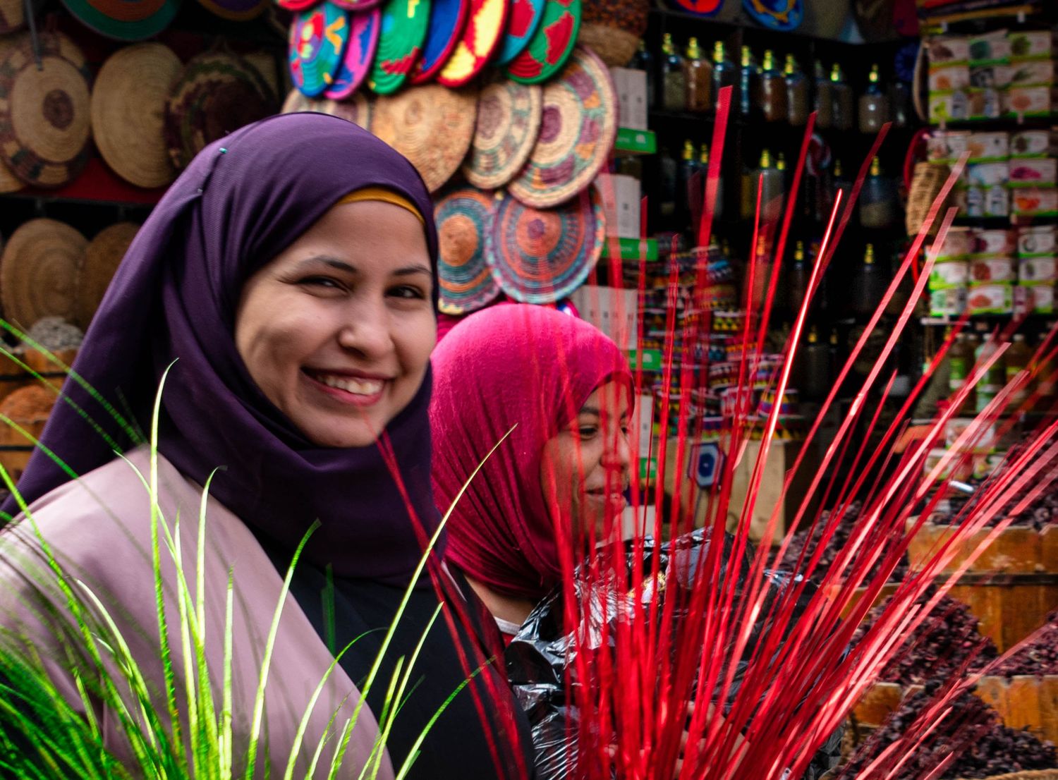 Two women wearing colorful hijabs smile at a vibrant market stall displaying decorative plates and red ornaments.