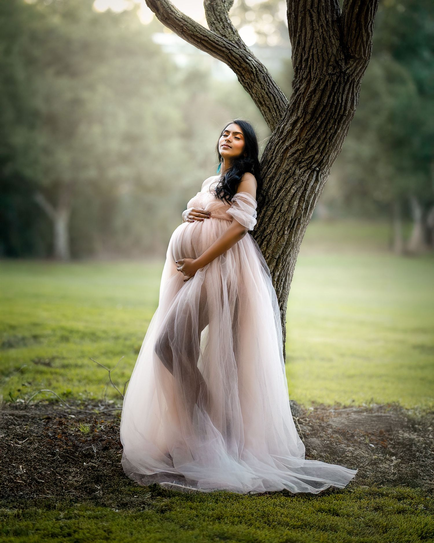 A figure in a flowing pink tulle dress poses gracefully against a tree trunk in a dreamy outdoor setting.