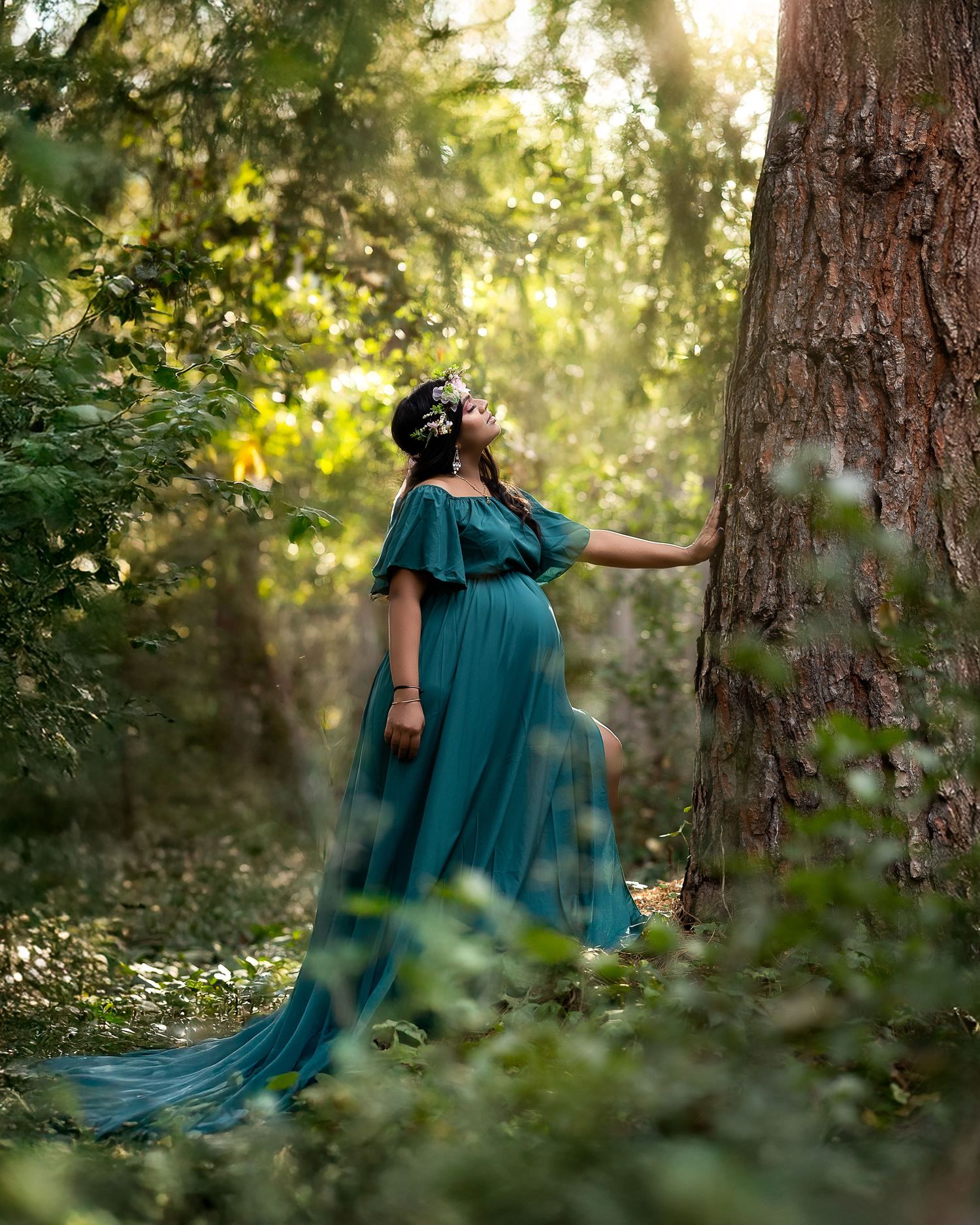 A figure in a flowing teal maternity gown stands dreamily in a sunlit forest setting with golden bokeh effects.