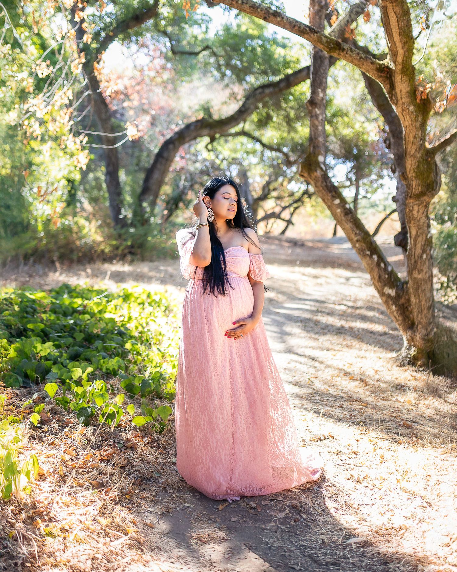 A person in a flowing pink dress poses gracefully among trees on a sunlit forest path during golden hour.
