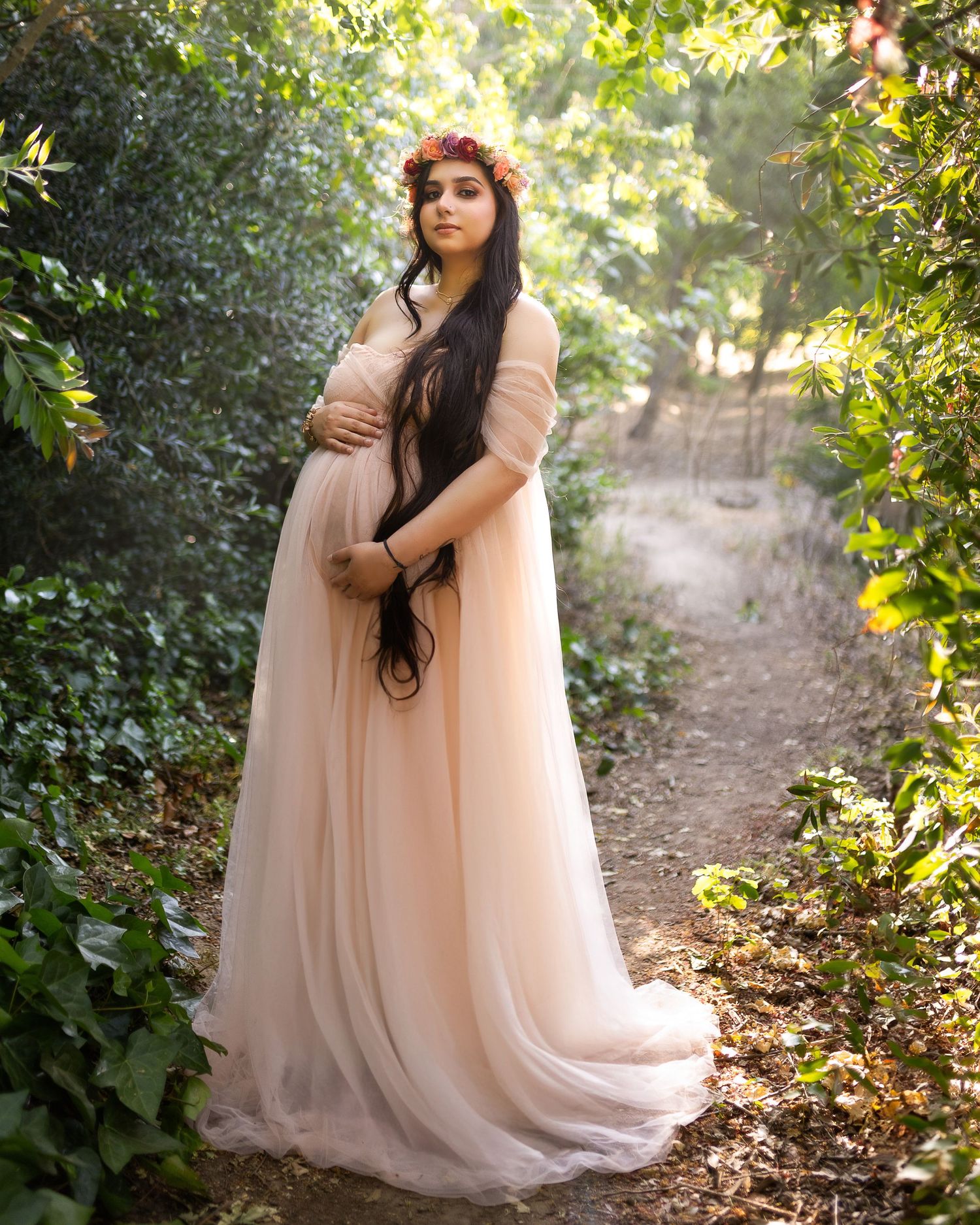 A figure in a flowing blush pink gown poses gracefully among lush greenery in a sunlit forest setting.