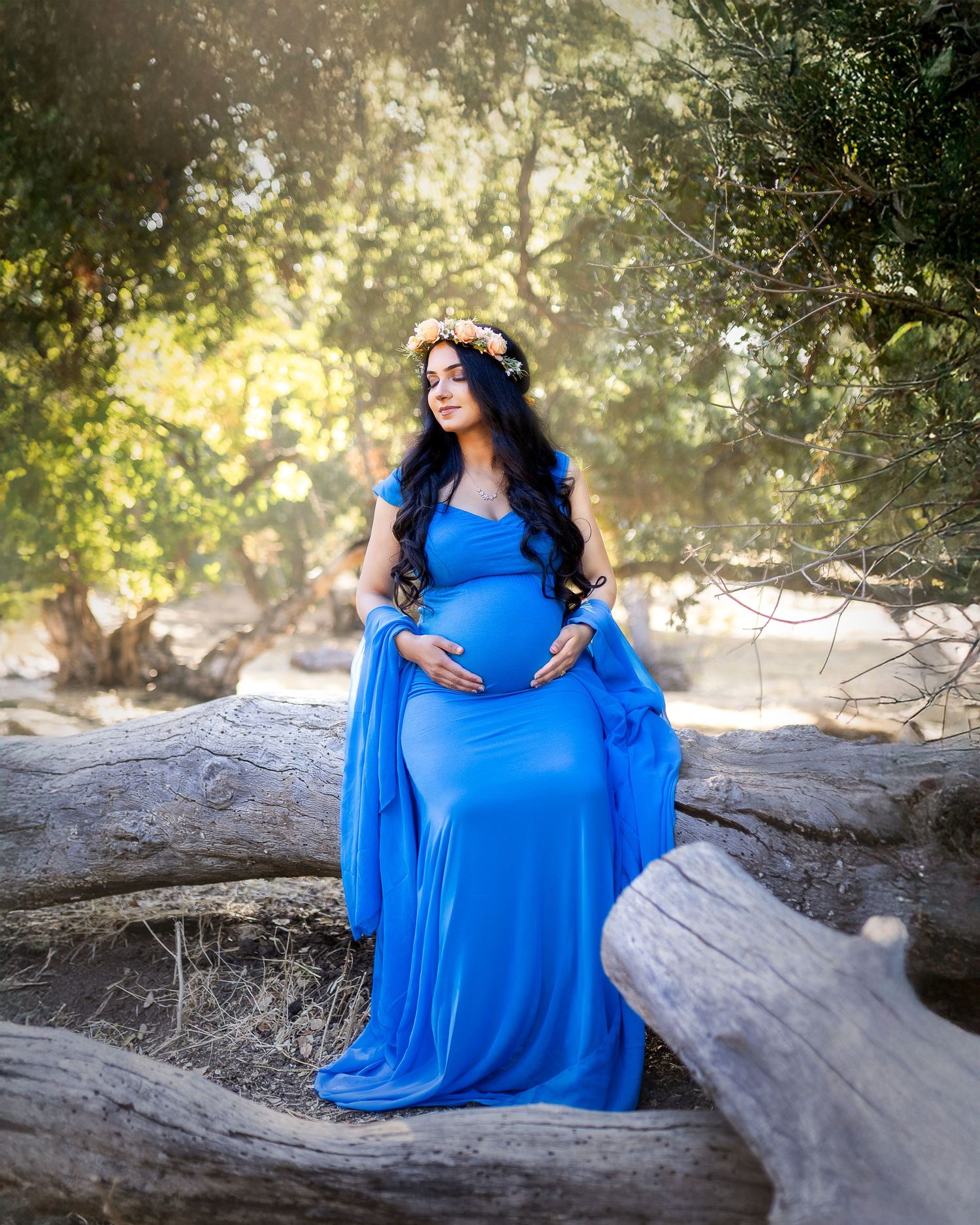 A woman in a flowing bright blue maternity dress poses gracefully on a fallen tree trunk in dappled sunlight.