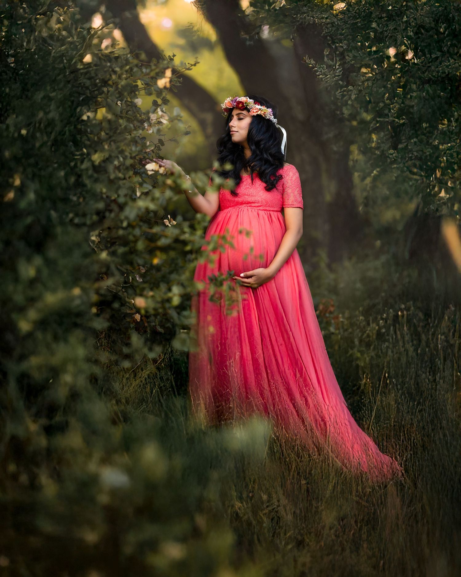 A person in a flowing coral pink gown stands gracefully in a dreamy outdoor setting during golden hour.