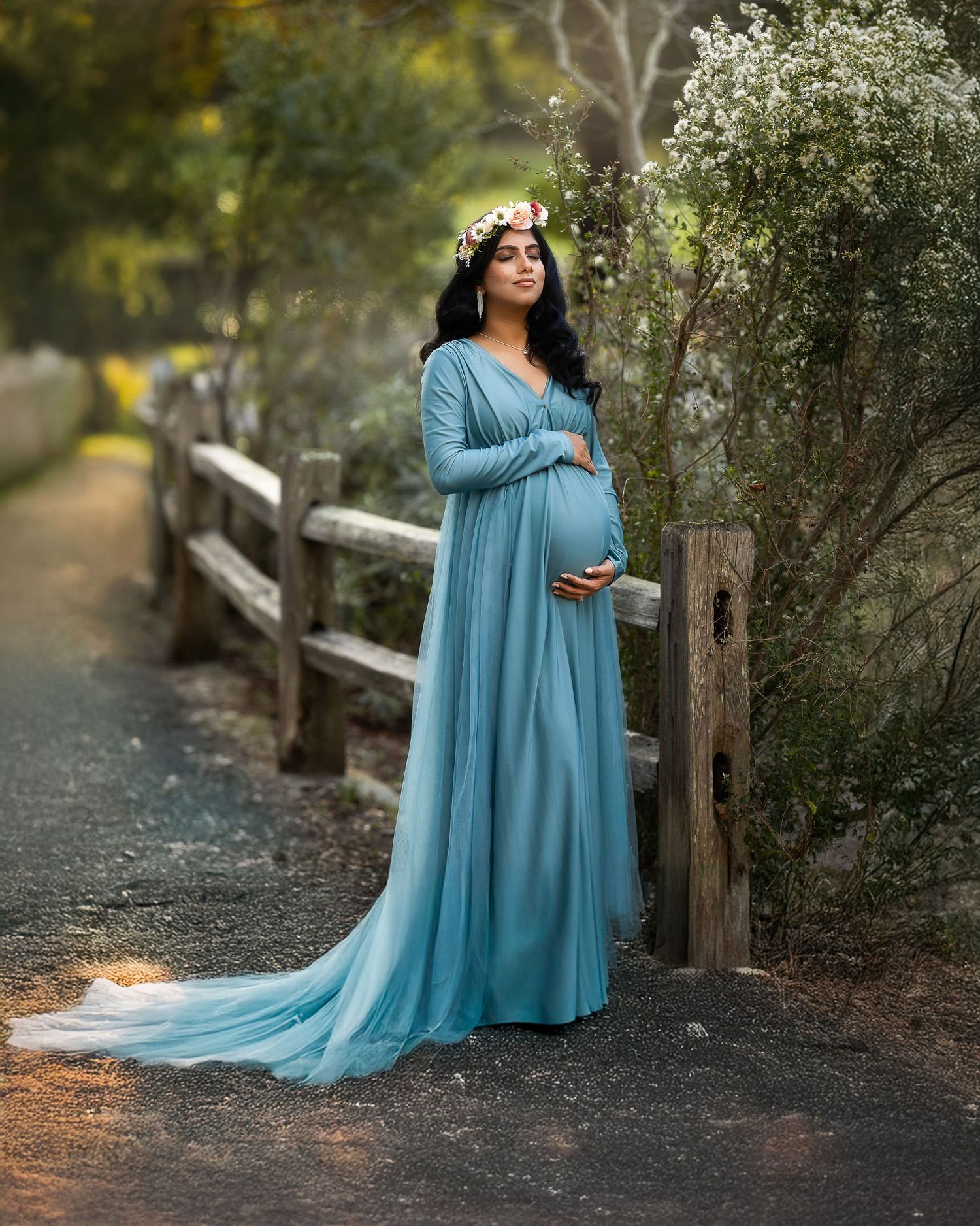 A flowing teal maternity gown with a long train photographed in a wooded outdoor setting during golden hour.
